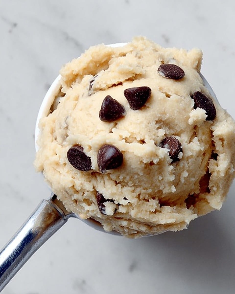 The image shows a close view of a single, thick scoop of light beige dough with a soft, lumpy texture, dotted with several dark brown chocolate chips sitting on top. The dough is in a small, white container with a shiny silver spoon resting against it. The background is a plain white marbled texture. The photo taken with an iphone --ar 4:5 --v 7