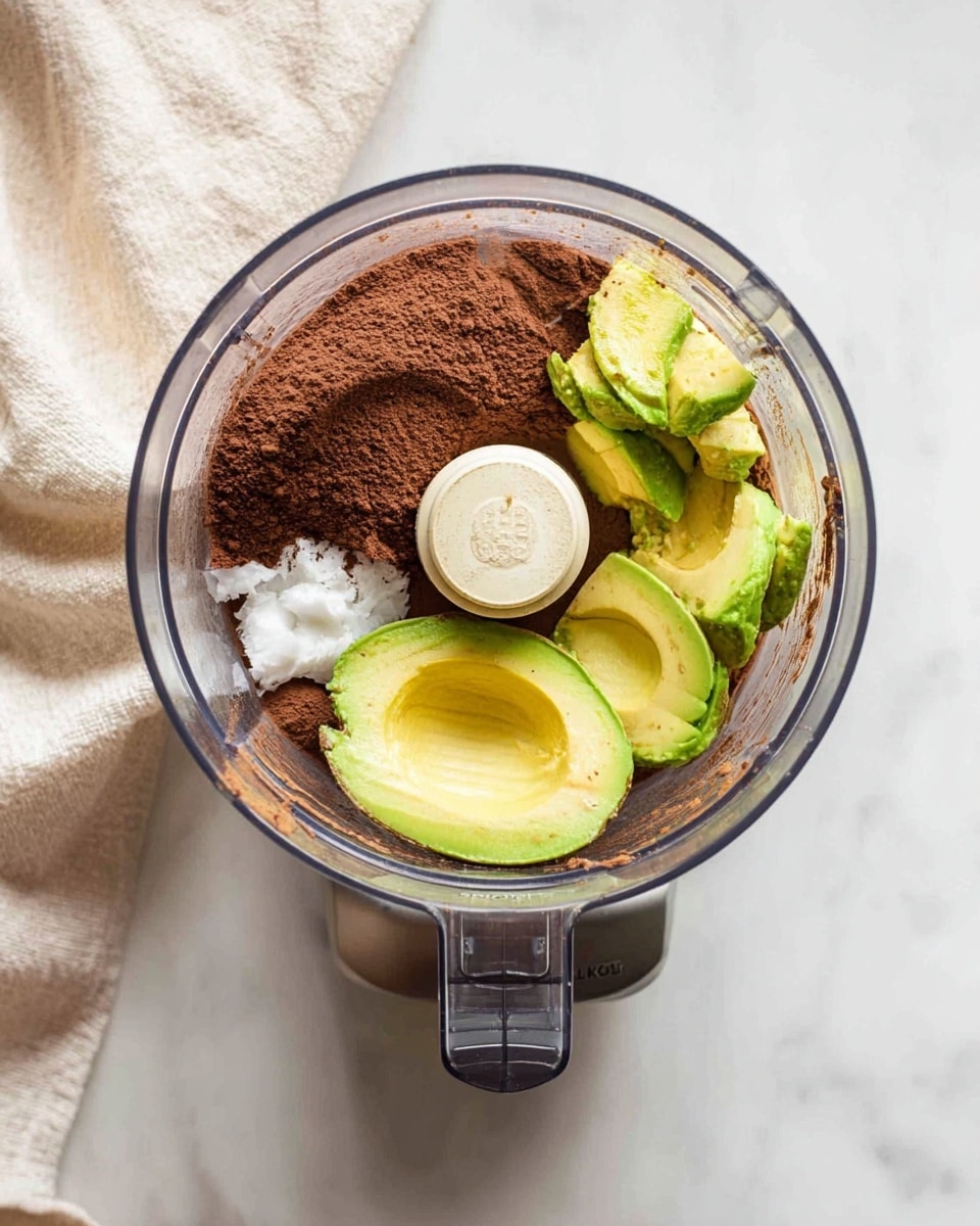 A clear food processor bowl is filled with several ingredients before blending: three avocado halves with green and yellow tones are positioned on the right side, next to a large portion of dark brown cocoa powder spread on the left side. Two small dollops of bright white coconut oil are on top of the cocoa powder near the center. The base of the processor is silver and gray, sitting on a white marbled surface with a light beige cloth partially visible to the left. photo taken with an iphone --ar 4:5 --v 7