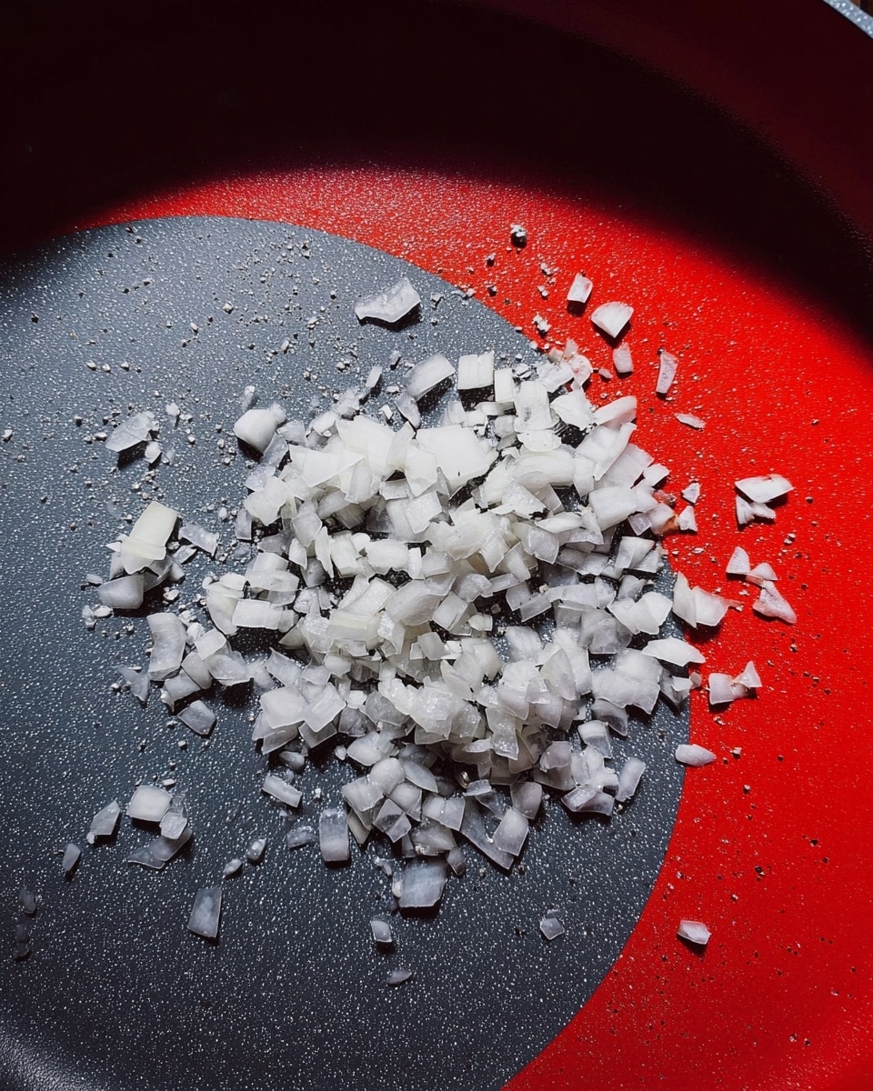 A close-up of a grey pan with small pieces of chopped white onion spread inside. The onions have a slightly wet, shiny texture and are scattered unevenly across the pan’s smooth surface. The grey pan contrasts with the bright red area surrounding it. Photo taken with an iphone --ar 4:5 --v 7