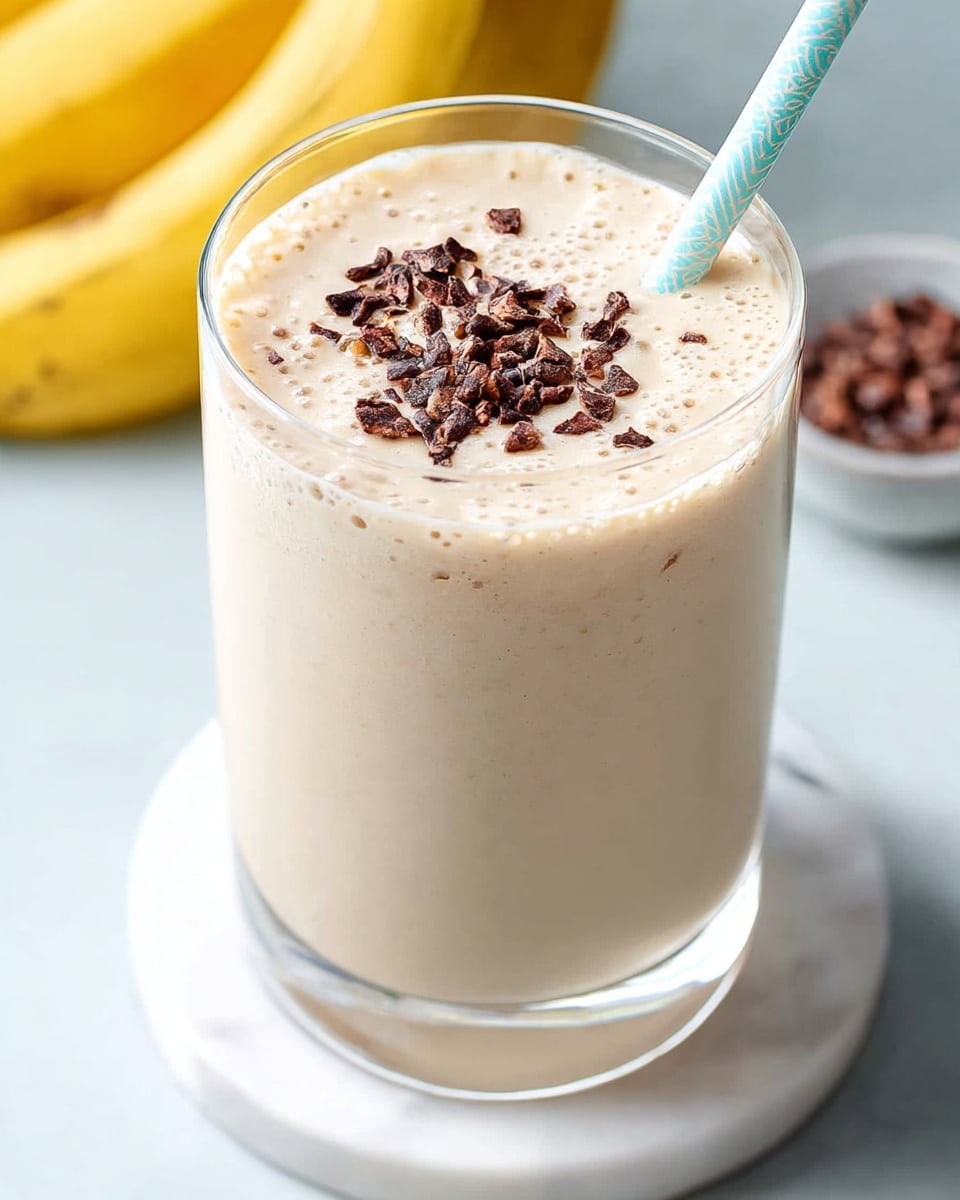 A clear glass filled with a thick, creamy light beige smoothie topped with small dark brown cacao nibs scattered on the surface, a light blue and white patterned straw leaning against the side, the glass placed on a white round coaster with a smooth white marbled surface underneath, and ripe yellow bananas blurred in the background, photo taken with an iphone --ar 4:5 --v 7