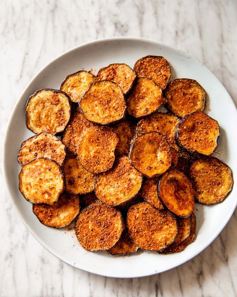 A white plate is filled with many round, thin slices of eggplant that are golden brown and slightly dark around the edges, showing a crispy texture. The slices overlap each other so the plate looks full, and each piece is covered with a light orange spice powder on top, adding texture and color contrast. The plate sits on a white marbled surface that gives a clean and bright look. photo taken with an iphone --ar 4:5 --v 7