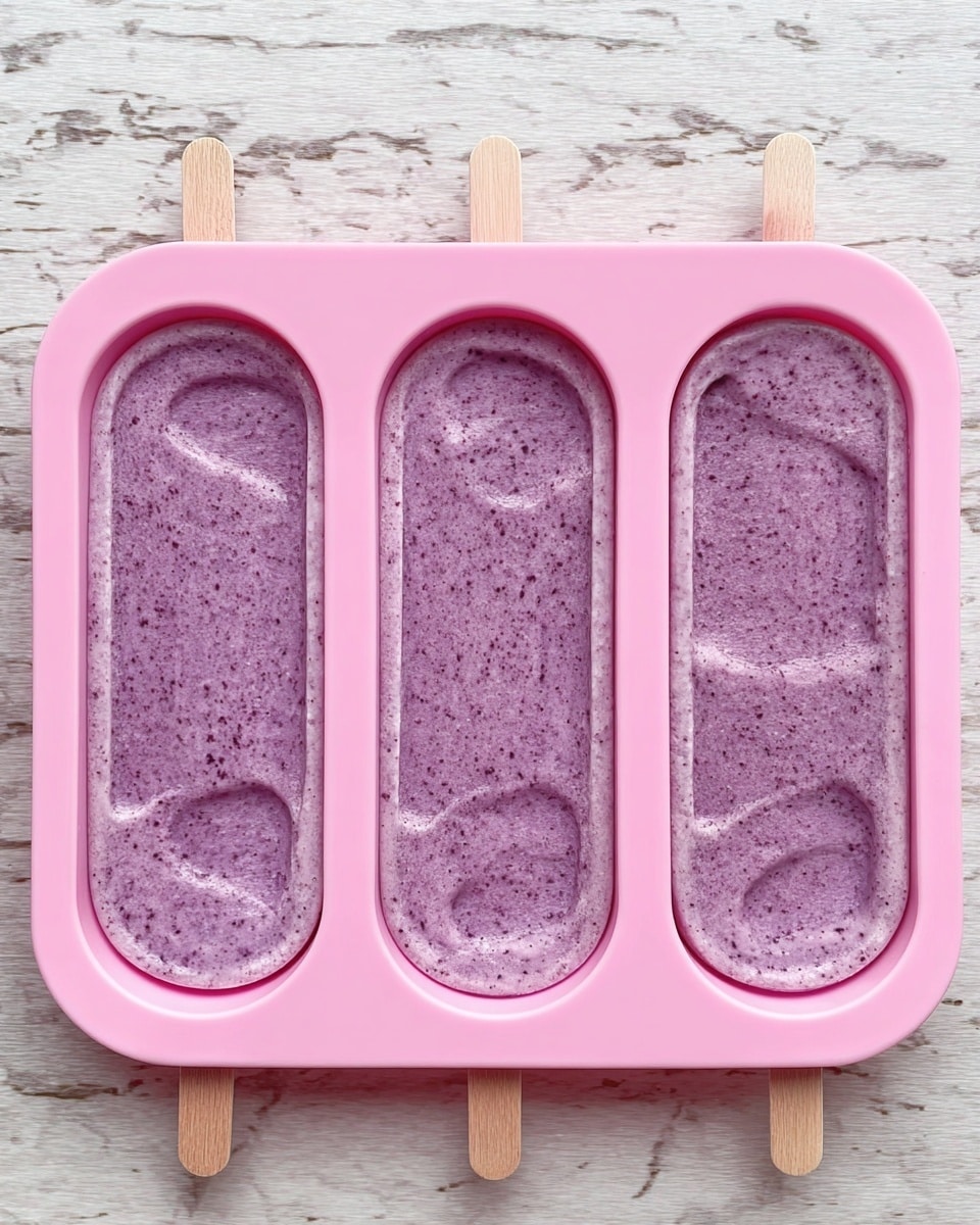 In the image, there is a pink silicone mold with three oval sections laid on a white marbled surface. Each section is filled with a purple mixture that has a creamy, slightly textured look, visible with tiny darker specks throughout. On the right side, wooden sticks are inserted into the mixture in each mold slot, showing the preparation for popsicles. The pink mold contrasts with the white marbled background and the natural wood sticks, emphasizing the purple mixture inside. The scene is simple and clean, focusing on the mold and its contents. photo taken with an iphone --ar 4:5 --v 7
