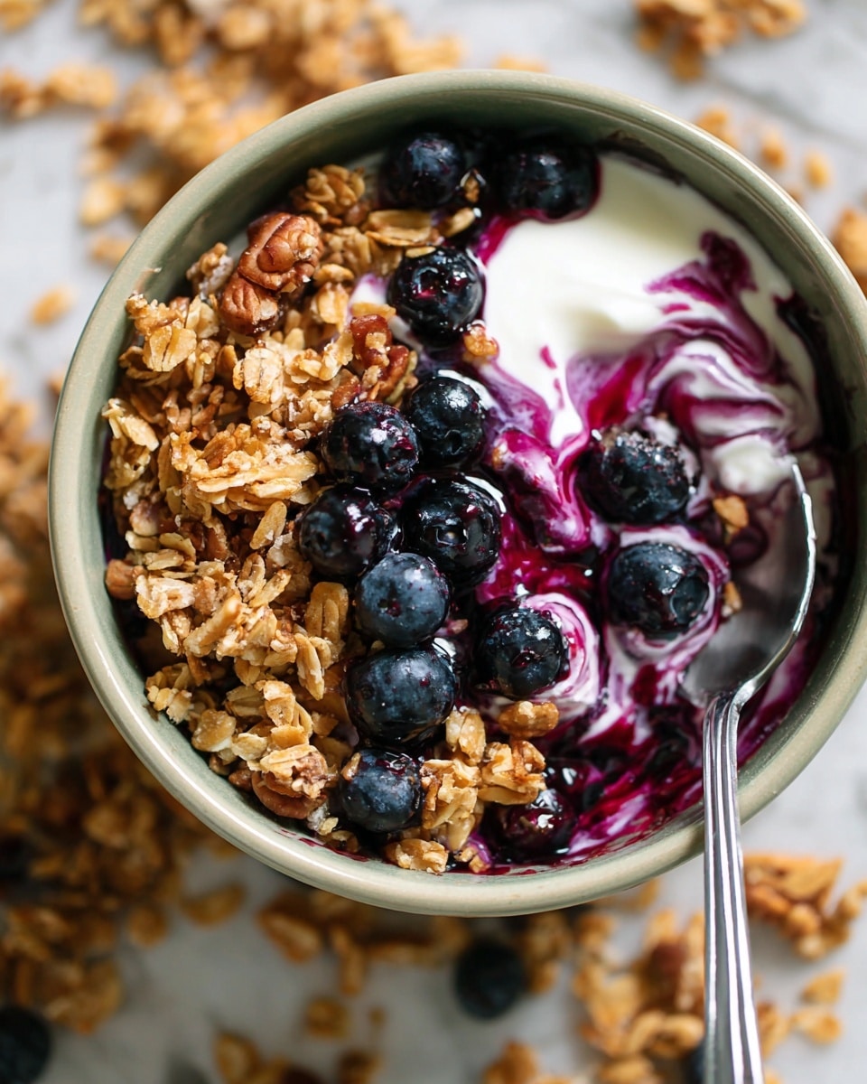 A close-up view of a white bowl filled with three main layers: a creamy white yogurt swirled with deep purple blueberry sauce at the bottom, topped with a layer of golden brown granola mixed with oats and nuts, and scattered fresh, plump blueberries resting on top, some partially covered in yogurt. A shiny metal spoon rests on the right side inside the bowl, holding some yogurt and granola. The bowl is placed on a white marbled surface with loose granola scattered around. Photo taken with an iphone --ar 4:5 --v 7
