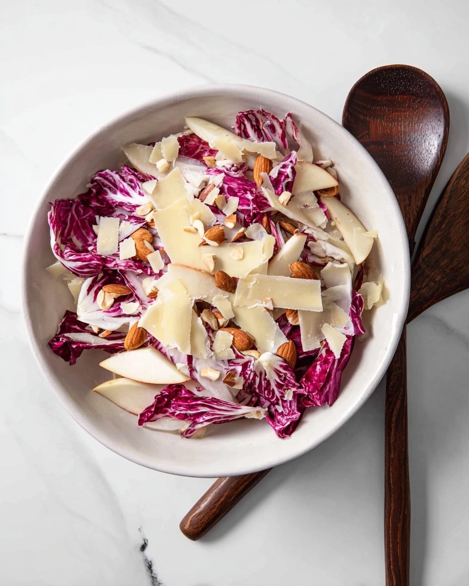 A white bowl filled with a salad showing three main layers: the bottom layer is light cream-colored thinly sliced apple pieces, the middle layer has red and white radicchio leaves with a crisp and slightly crinkled texture, and the top layer is thin pale yellow cheese shavings scattered unevenly, along with chopped almonds sprinkled over the salad. The bowl is placed on a white marbled surface with a dark wooden serving spoon next to it. Photo taken with an iphone --ar 4:5 --v 7