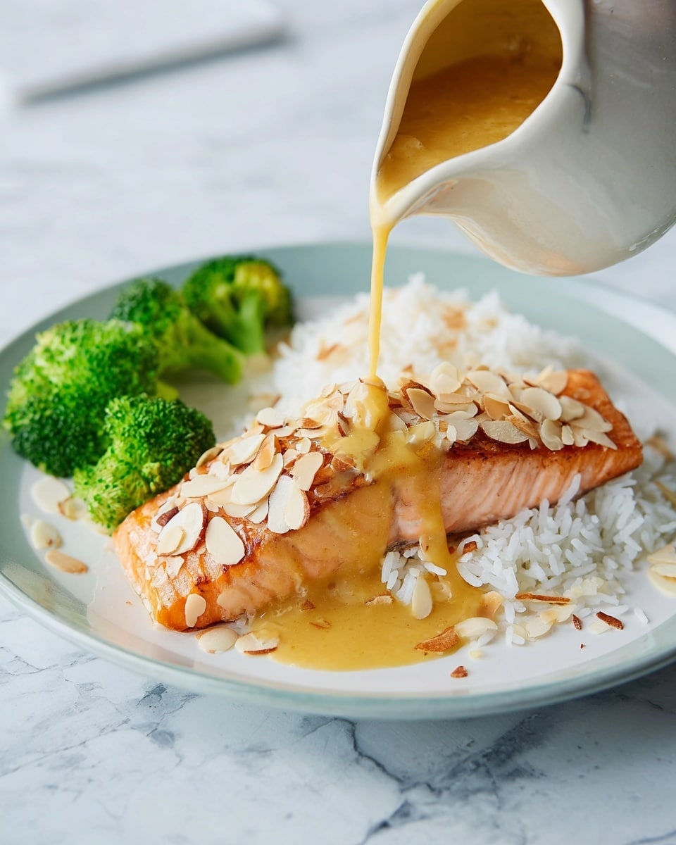 A white plate shows a cooked salmon fillet at the front covered with thin toasted almond slices, with a thick golden sauce being poured over it from a white sauce pitcher; behind the salmon, there is a serving of white rice, and three bright green broccoli florets are placed near the edges of the rice and salmon. The surface under the plate is a white marbled texture. photo taken with an iphone --ar 4:5 --v 7
