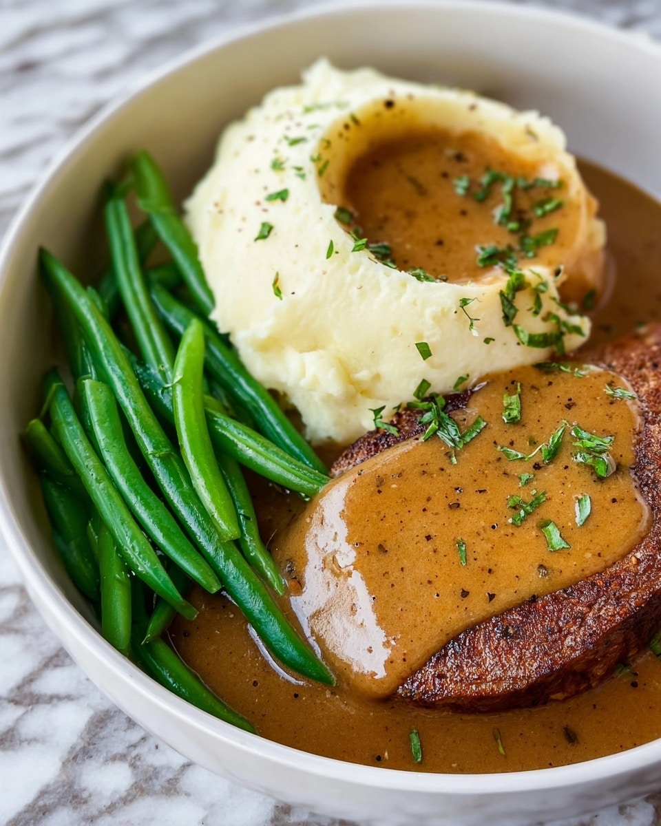 A white bowl holds three main layers: on one side, bright green string beans arranged close together, showing a smooth, slightly shiny texture; next to them, a round mound of creamy, white mashed potatoes with a soft, fluffy texture and a small well on top filled with golden brown gravy speckled with black pepper; beside the mashed potatoes, a thick, roasted piece of cooked meat with a slightly browned, textured surface, also covered by the same rich brown gravy spreading gently across the bowl and sprinkled with small bits of chopped fresh green herbs. The whole dish is placed on a white marbled texture surface. Photo taken with an iphone --ar 4:5 --v 7