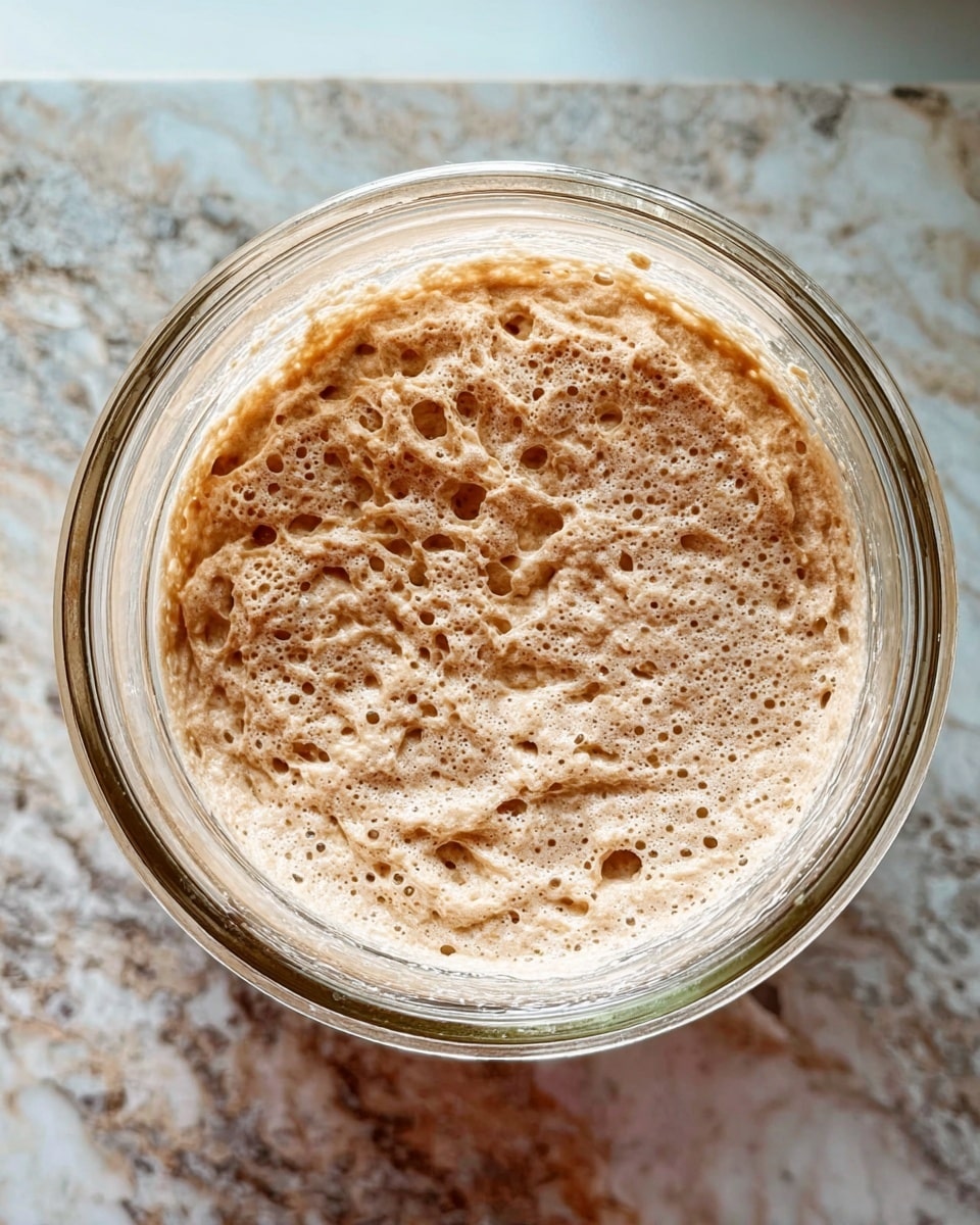 A close-up top view of a clear glass jar filled with a light brown dough that has a bubbly and slightly sticky texture. The dough fills most of the jar’s inside surface, showing some uneven, spongy holes all over. The jar is sitting on a white marbled surface with gray and brown veins, providing a soft contrast to the dough. Photo taken with an iphone --ar 4:5 --v 7