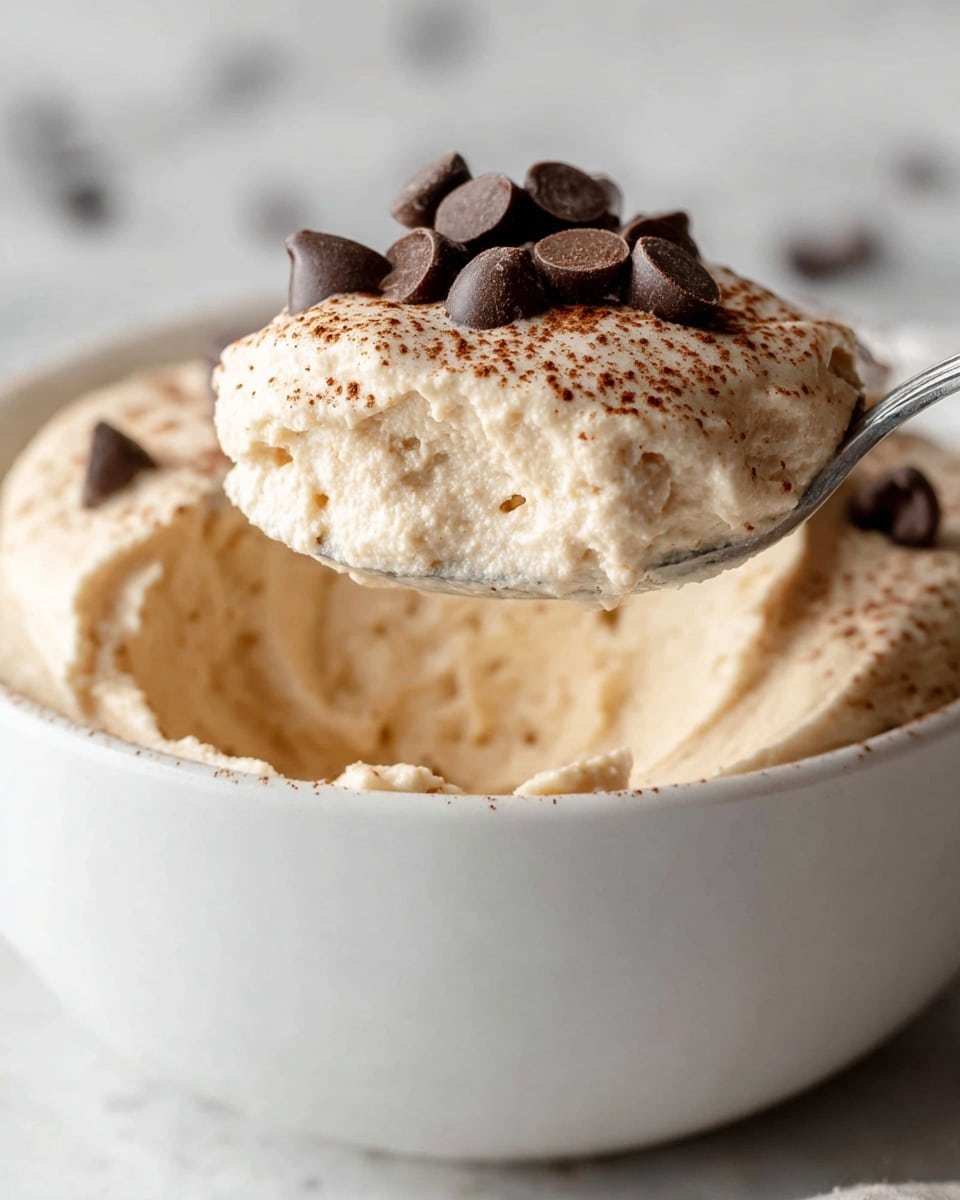 A close-up of a white bowl filled with creamy, light beige mousse. A spoon lifts a scoop of the mousse, showing its soft, fluffy texture with a smooth surface lightly dusted with cocoa powder. On top of the mousse scoop, there are several dark brown chocolate chips scattered, adding contrast and texture. The background is a white marbled texture, and the photo is clear and bright. photo taken with an iphone --ar 4:5 --v 7