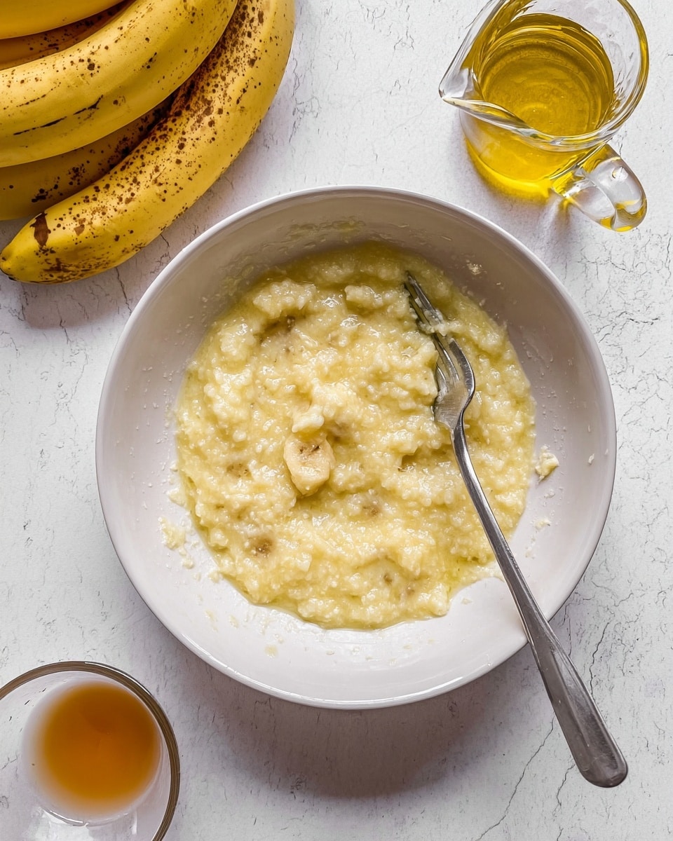 A white bowl contains a mushy, light yellow mixture with small lumps, likely mashed banana, with a silver fork resting in it. At the top left, part of a bunch of ripe bananas with brown spots is visible. To the right of the bowl, there is a small clear glass pitcher filled with golden oil and a small glass dish with a light brown liquid. The background surface is a white marbled texture. Photo taken with an iphone --ar 4:5 --v 7