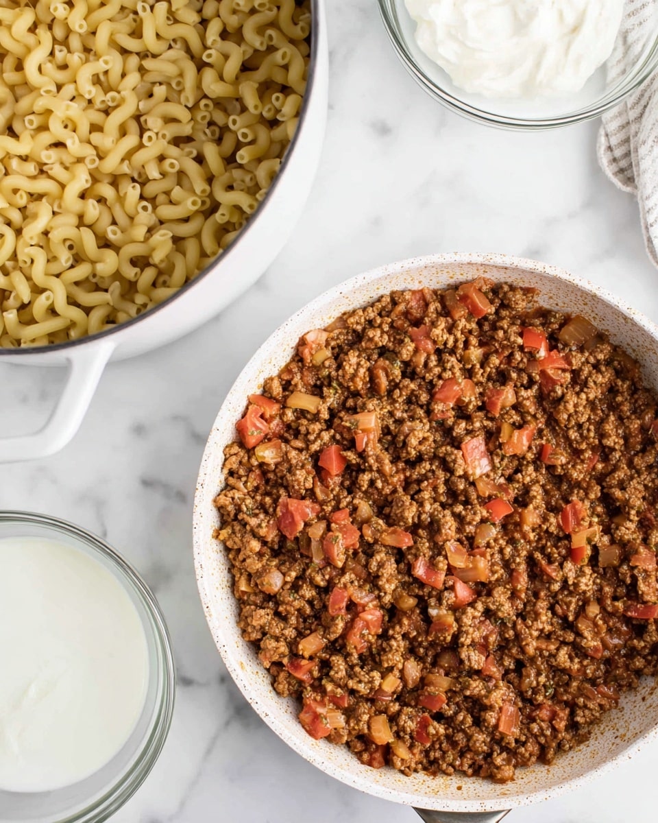 The image shows a close-up of two pans on a white marbled surface. The pan at the bottom is a white skillet filled with cooked ground meat mixed with diced tomatoes and small pieces of onion and herbs, displaying a rich brown and red color with a slightly chunky texture. Above it to the left, there is a white pot filled with cooked elbow macaroni, showing a pale yellow color and smooth curved shapes. To the right, there are two clear glass bowls containing white creamy substances. The image has a bright, clean look with the white marbled background and simple cookware. Photo taken with an iphone --ar 4:5 --v 7