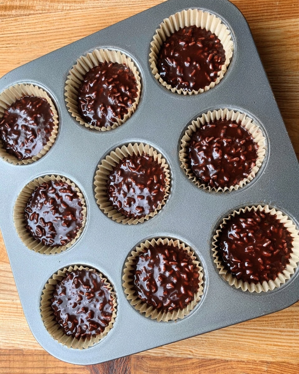 A dark rich chocolate mixture with shiny texture and small rice crisps is placed in six beige paper liners inside a gray metal muffin tray. The chocolate mix fills each liner unevenly but almost to the top, showing the texture of rice crisps throughout. The tray is set on a light wooden surface with a natural grain pattern. Photo taken with an iphone --ar 4:5 --v 7