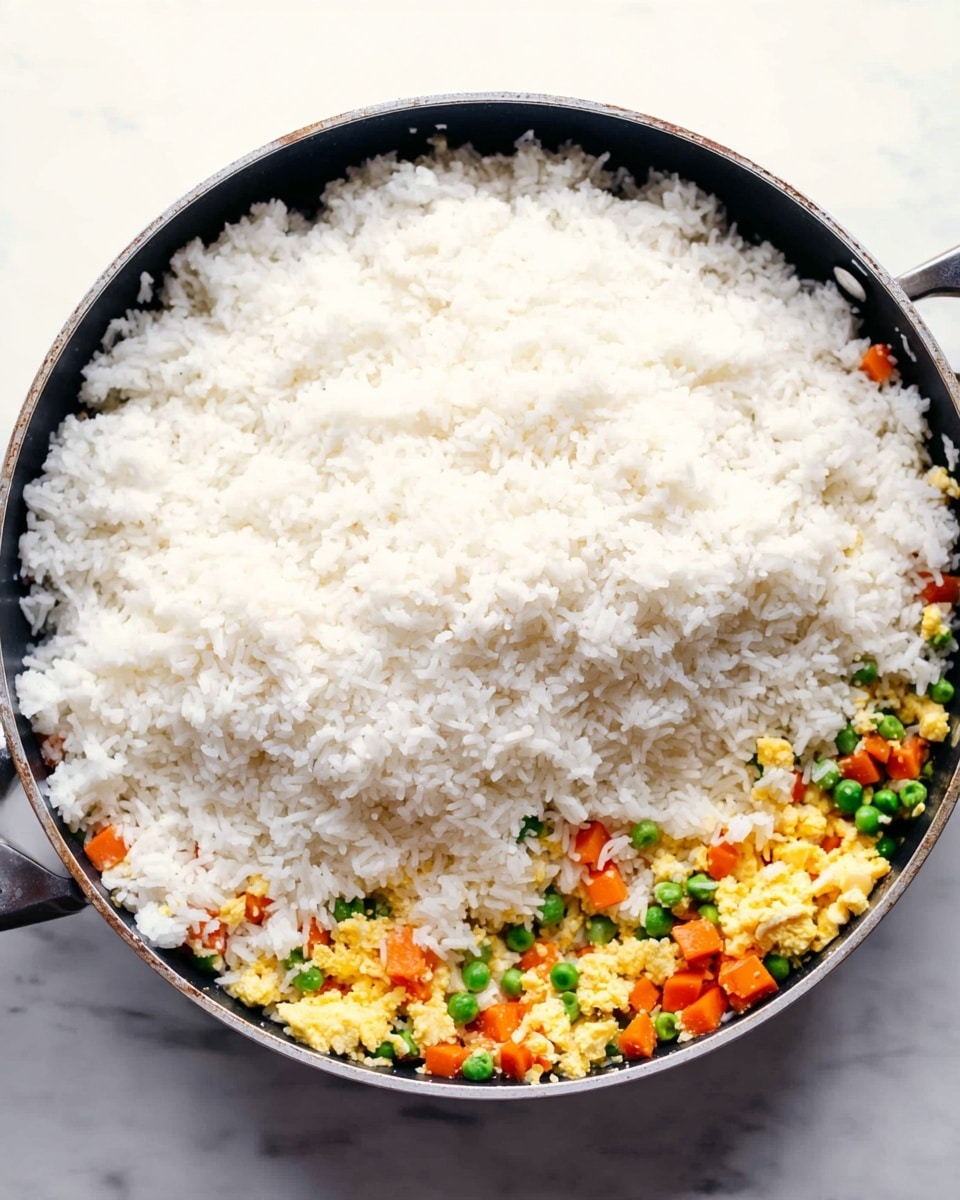 A large black pan filled mostly with a thick layer of white cooked rice covering the top and center. Underneath the rice, there is a colorful layer mostly visible on the lower right side, made of small diced orange carrots, bright green peas, and yellow scrambled egg pieces mixed with some more white rice grains. The pan is placed on a white marbled surface. Photo taken with an iphone --ar 4:5 --v 7