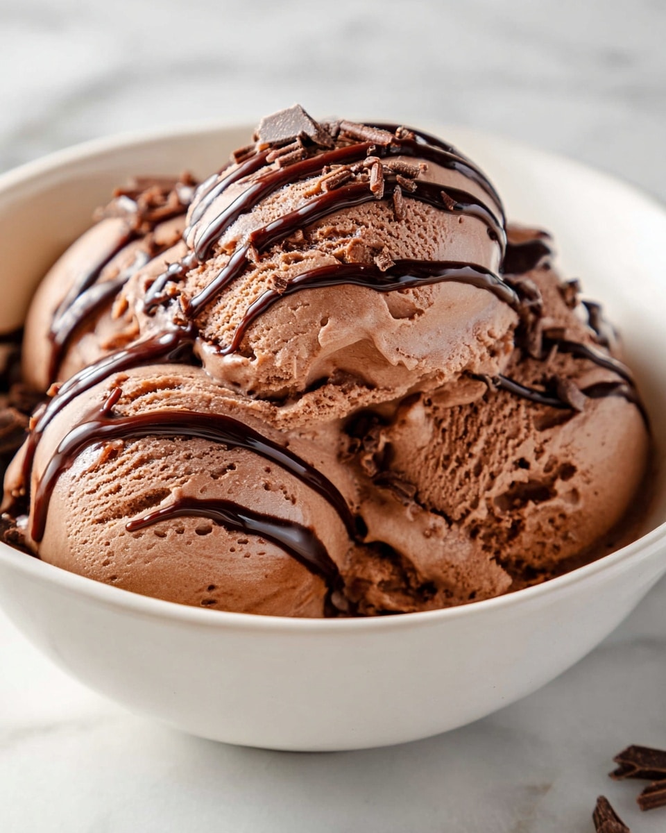 A white bowl filled with three scoops of smooth chocolate ice cream, each scoop showing a creamy brown color with small air holes and little chunks inside. Thick lines of dark chocolate sauce are drizzled across the top of the scoops, adding glossy dark stripes that contrast with the lighter brown ice cream. Small, thin pieces of chocolate sprinkles are scattered over the ice cream, adding texture. The bowl sits on a white marbled surface with soft natural light that highlights the ice cream’s creamy texture. photo taken with an iphone --ar 4:5 --v 7