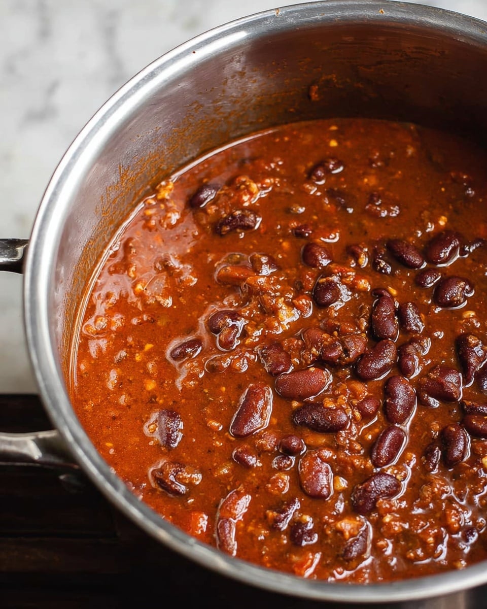 A close-up view of a pot filled with thick, homemade chili showing one layer of rich reddish-brown sauce mixed with whole and soft beans. The beans are dark brown and shiny, sitting in a glossy, slightly chunky sauce with visible small bits of spices and vegetables. The shiny metal inside of the pot has measurement marks along the side. The pot rests on a white marbled surface. Photo taken with an iphone --ar 4:5 --v 7