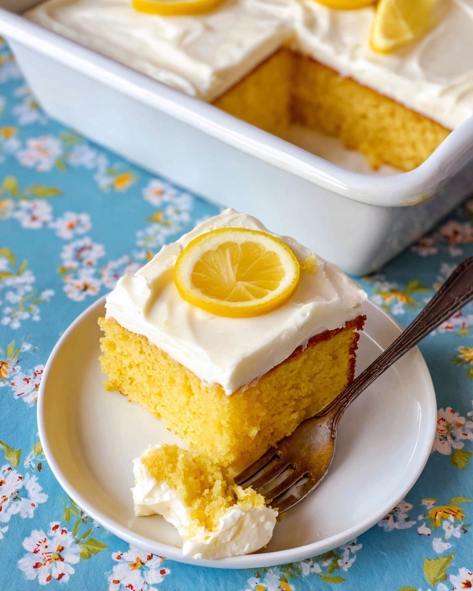 A piece of yellow cake with two layers is topped with smooth white frosting and a thin slice of yellow lemon sitting in the middle on a white plate. Next to the cake piece is a fork with some frosting on it. The plate is placed on a table with a blue floral tablecloth. In the background, there is a white baking pan with the rest of the cake that has a piece missing. The surface is a white marbled texture photo taken with an iphone --ar 4:5 --v 7