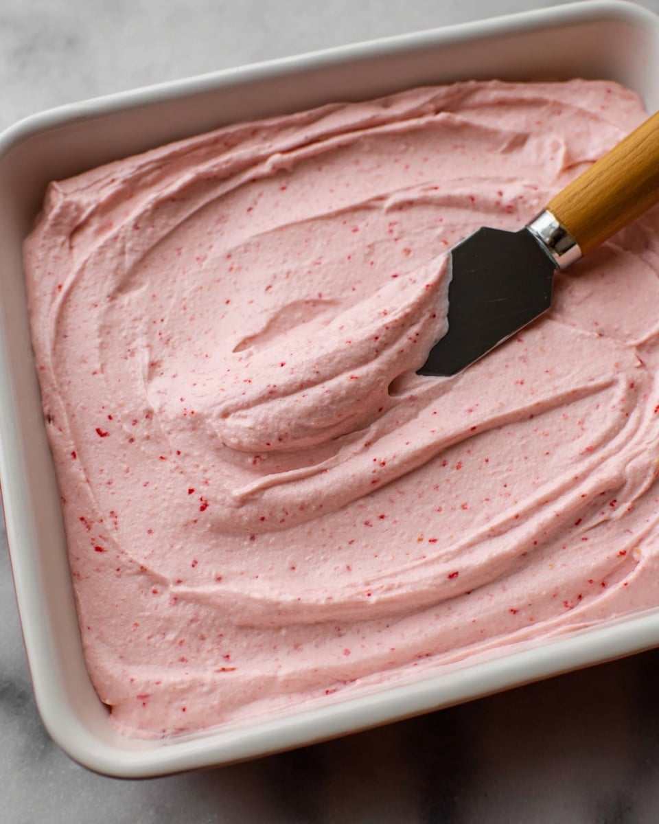 A close-up view of a white rectangular dish filled with a thick, smooth, and creamy light pink spread that has tiny red specks mixed throughout. The spread is evenly layered, covering the entire dish with some soft swirls and ridges on the surface. A spatula with a black blade and wooden handle rests in the spread, with some of the pink mixture on its blade. The dish is placed on a white marbled surface. photo taken with an iphone --ar 4:5 --v 7