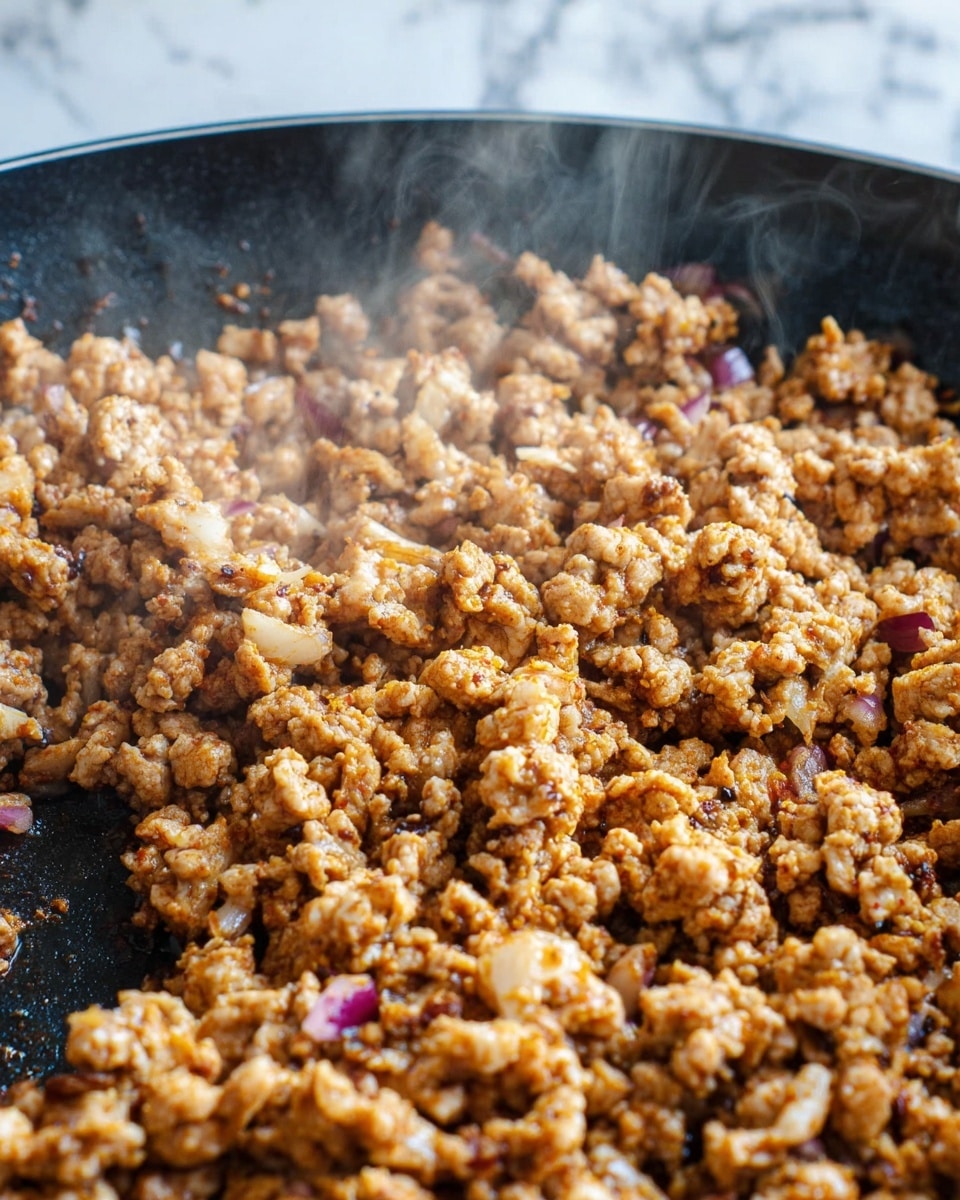 The image shows a close-up of cooked ground meat mixed with small pieces of red onion in a black pan. The meat is a light brown color with a slightly orange tint from seasoning, and the small onion bits add specks of purple throughout. The texture of the cooked meat looks crumbly and well-cooked, with visible steam rising, indicating heat. The black pan contrasts with the warm tones of the food. The background is a white marbled texture. Photo taken with an iphone --ar 4:5 --v 7