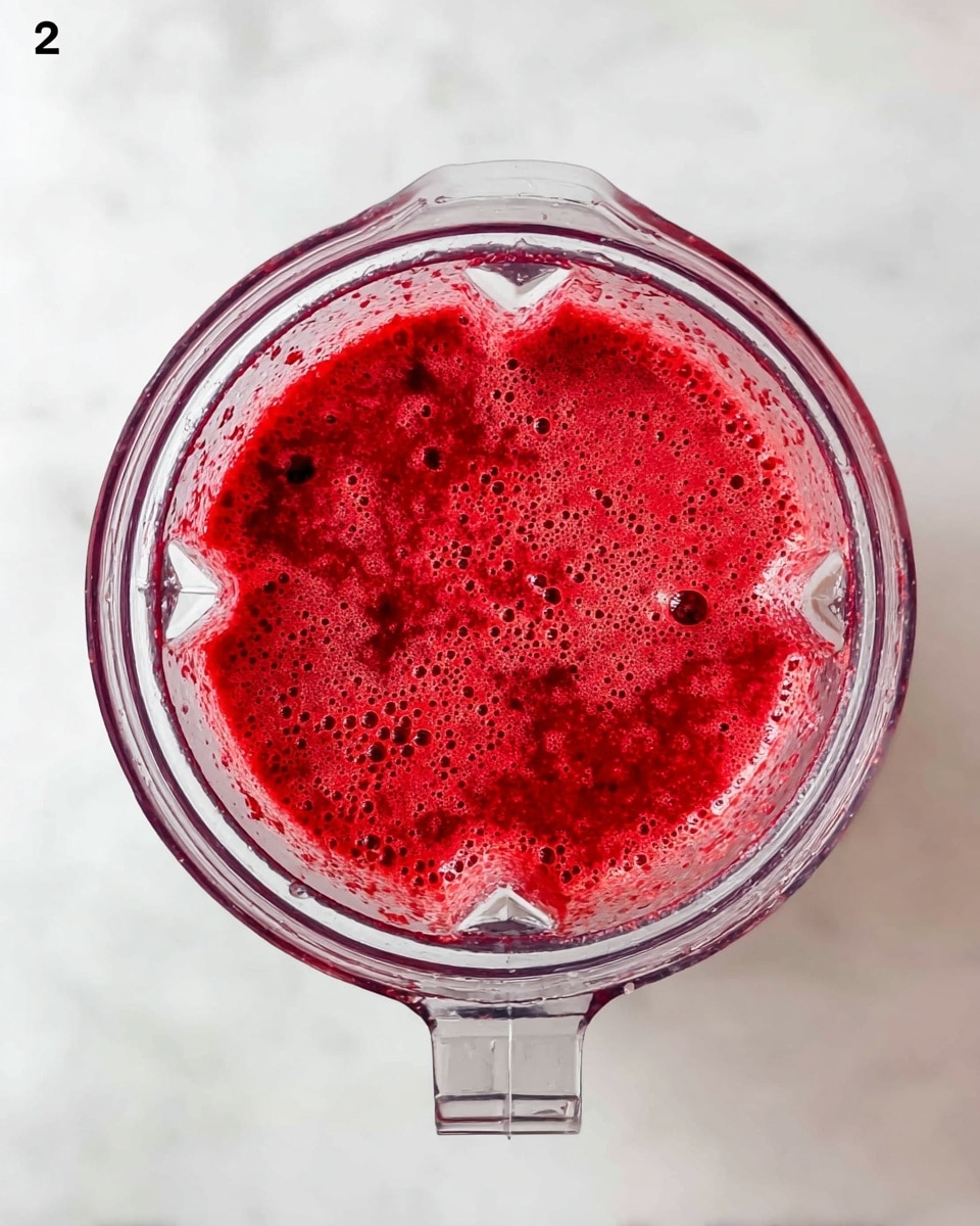The image shows a clear blender jar viewed from above, filled with a bright red smoothie mixture. The smoothie has a foamy texture with small bubbles and specks of darker red scattered throughout. The blender jar is placed on a white marbled surface, enhancing the vibrant color of the smoothie inside. Photo taken with an iphone --ar 4:5 --v 7