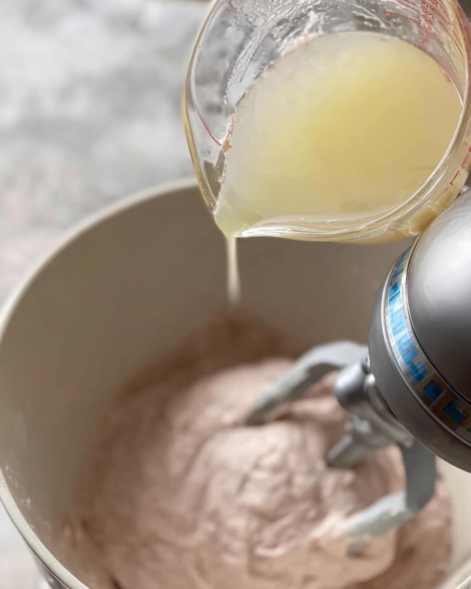 A close-up view shows a white mixer bowl at the bottom filled with a thick, light pink mixture being mixed by a silver electric hand mixer with beaters inside the bowl. Above the bowl, a glass measuring cup with a light yellow liquid is being poured into the bowl, with the liquid forming a thin stream between the cup and the bowl. The scene is set on a surface with a white marbled texture, softly blurred in the background. Photo taken with an iphone --ar 4:5 --v 7