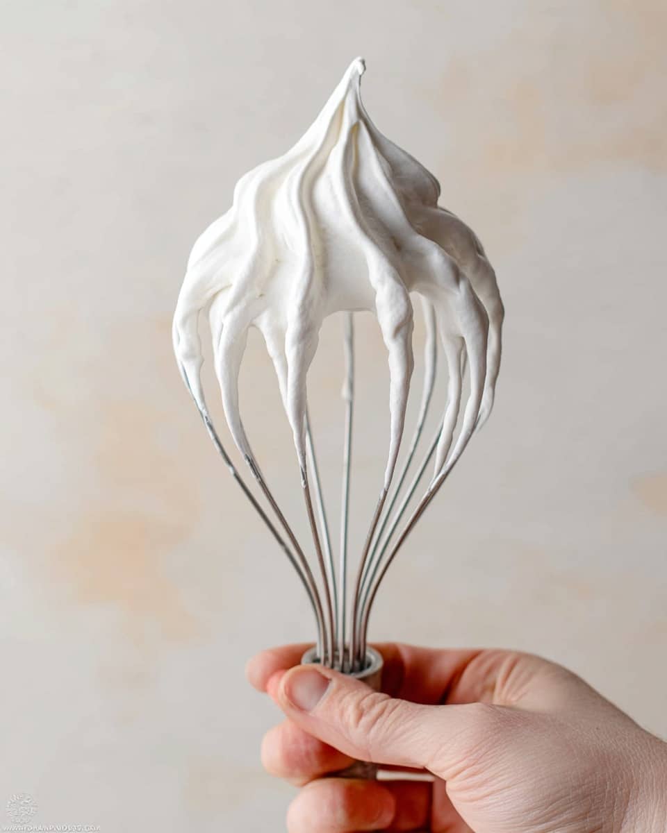 A close-up image of a metal whisk with a soft, white whipped cream or meringue clinging to its wires, forming a smooth peak at the top. The textures are fluffy and airy, with some strands hanging down from the wires. A woman's hand holds the bottom part of the whisk, with visible fingers gripping it gently. The background is a soft, neutral beige tone with a white marbled texture surface below. photo taken with an iphone --ar 4:5 --v 7