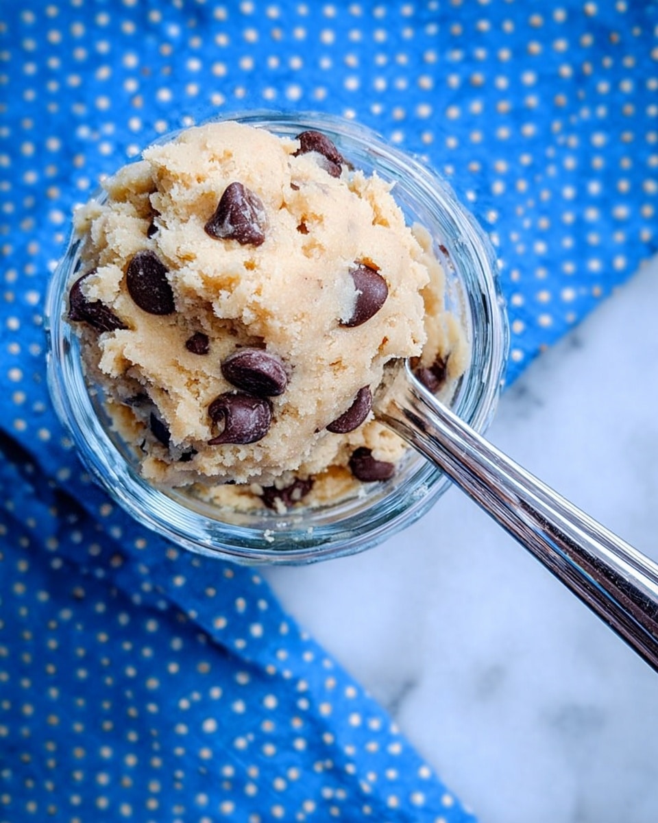 A small clear glass bowl filled to the top with light beige cookie dough mixed with large dark brown chocolate chips, some chips visible near the surface. A shiny metal spoon with a rounded end is placed inside the bowl and sticking out to the right side. The background shows a blue cloth with white dots, and the bowl sits on a white marbled texture surface photo taken with an iphone --ar 4:5 --v 7