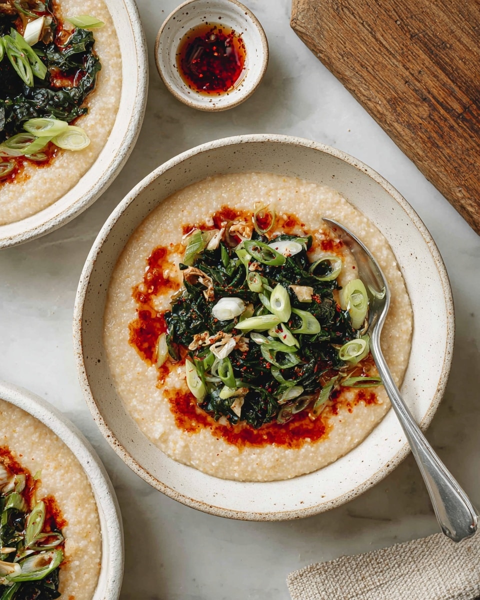 The image shows a close-up of three white speckled bowls filled with creamy beige grits as the bottom layer. On top, there is a drizzle of bright red chili oil spread unevenly, making a contrast with the pale grits. In the center of the front bowl, there is a pile of dark green sautéed greens, scattered with thinly sliced light green onions and small bits of translucent white onion. A silver spoon rests inside the front bowl, leaning towards the bottom right. To the upper left, a small white speckled bowl holds extra chili oil with a silver spoon inside. The dishes sit on a white marbled surface with a textured wooden board visible at the upper right. photo taken with an iphone --ar 4:5 --v 7