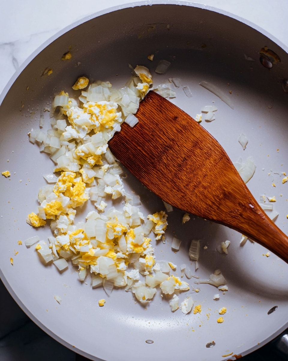 A close-up view of a white frying pan with small, unevenly cooked white onion pieces and bits of yellow scrambled egg scattered inside. The onions and eggs are spread primarily along the edges of the pan, with some pooling near the center. There is a wooden spatula resting inside the pan, its rich brown color contrasting with the pale ingredients and pan surface. The pan surface has a smooth, slightly shiny finish with a few droplets of oil visible. The background and surface seen under the pan are a white marbled texture. Photo taken with an iphone --ar 4:5 --v 7