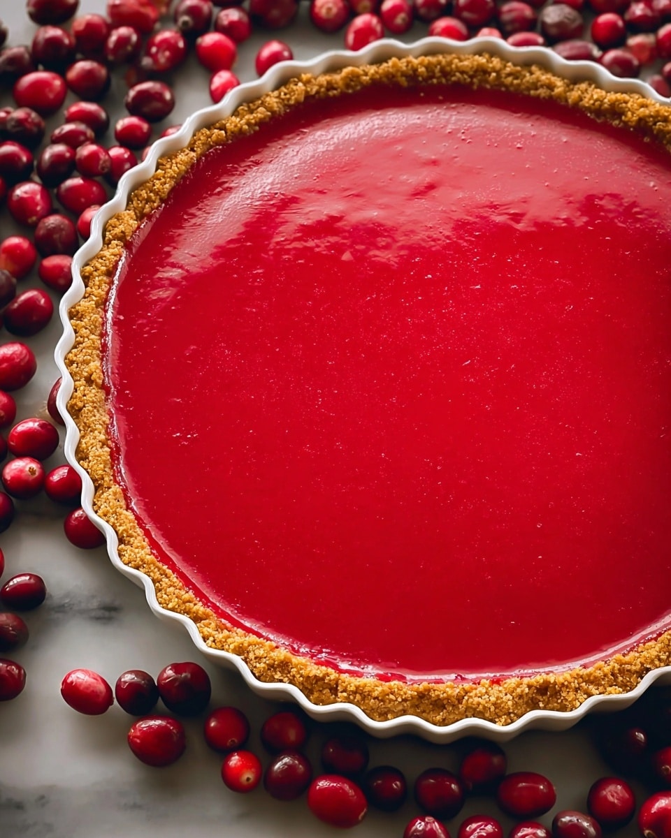 A round tart with two layers: the bottom layer is a light brown crumbly crust pressed into a fluted white tart pan, and the top layer is a smooth, shiny, bright red filling evenly spread to the edges. The tart sits on a white marbled surface scattered with fresh red cranberries around it. The colors are warm and rich, with a strong contrast between the bright red filling and the golden crust. Photo taken with an iphone --ar 4:5 --v 7