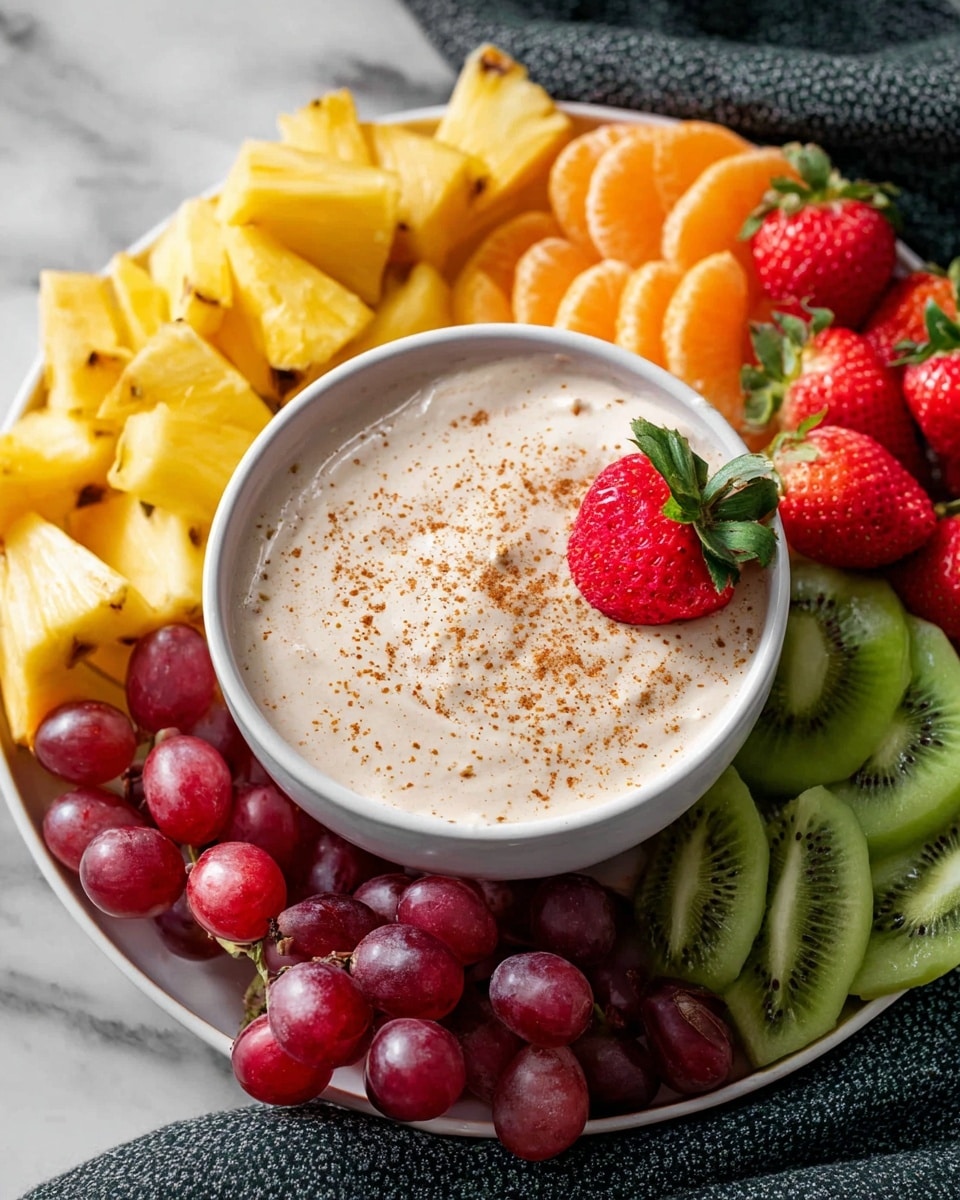 A white bowl in the center is filled with a creamy, light beige dip sprinkled with small brown spice specks. One red strawberry with green leaves is placed inside the dip at the edge. Around the bowl, there is a white plate holding an arrangement of fresh fruit: bright yellow pineapple chunks at the bottom left, thinly sliced green kiwi on the right, whole red strawberries at the top, peeled orange segments near the top left, and red grapes scattered at the bottom. The plate is set on a white marbled surface with a dark textured cloth in the background. photo taken with an iphone --ar 4:5 --v 7