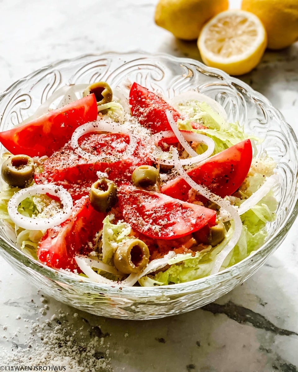 The dish is a fresh salad served in a clear glass bowl with decorative swirls. At the bottom, there is a layer of shredded pale green lettuce, topped with thin white onion slices spread evenly. On top of this, there are large, bright red tomato wedges scattered around, mixed with green olive halves stuffed with red pimento. The entire salad is sprinkled with a light dusting of white grated cheese and cracked black pepper. In the background, there are lemon halves on a white marbled surface. Photo taken with an iphone --ar 4:5 --v 7