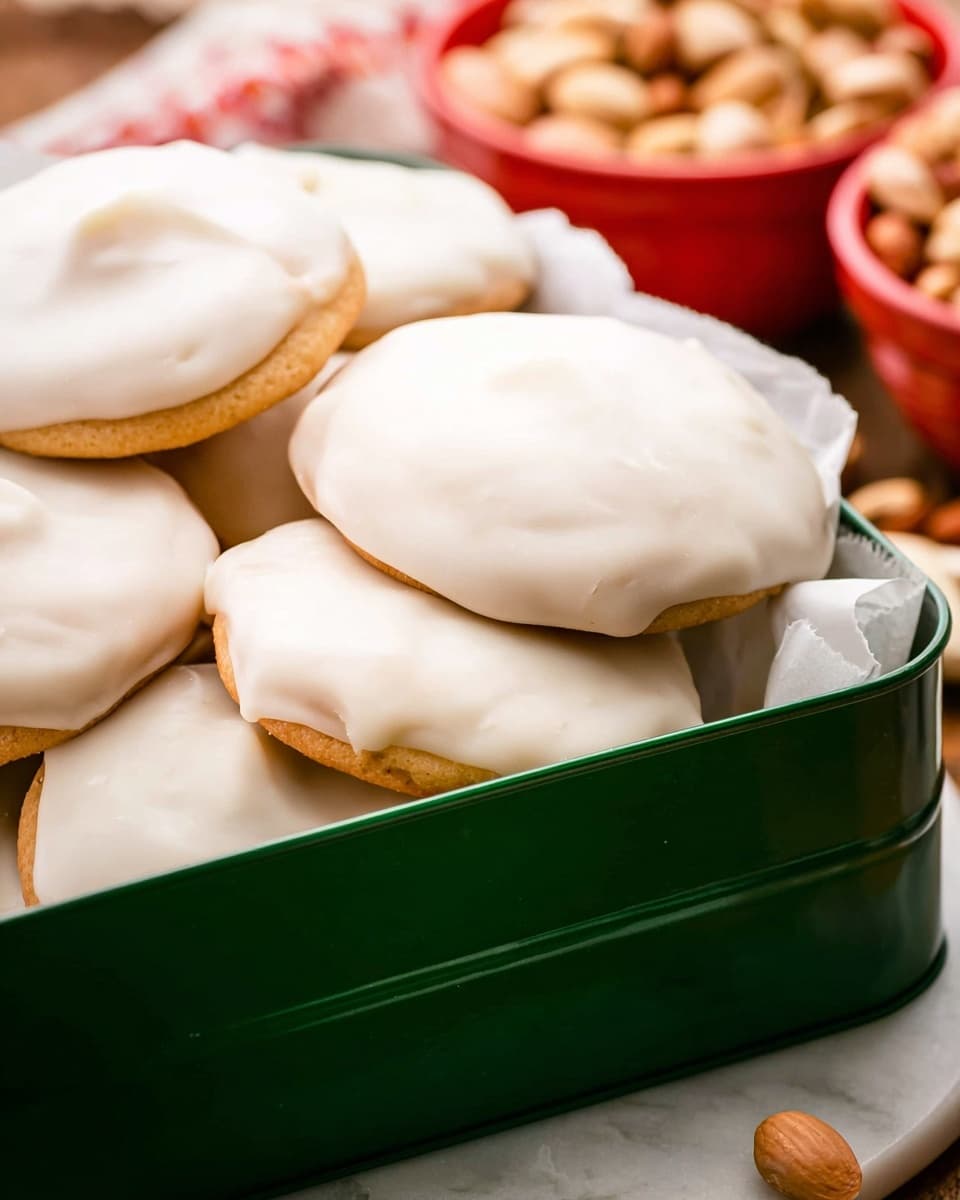 A green metal box filled with several round cookies stacked in layers, each cookie covered with a smooth, thick white icing that almost hides the golden-brown cookie base underneath. The cookies have an uneven, slightly textured icing surface that catches light softly. In the background, red bowls hold light tan nuts, and the scene rests on a white marbled texture. Photo taken with an iphone --ar 4:5 --v 7
