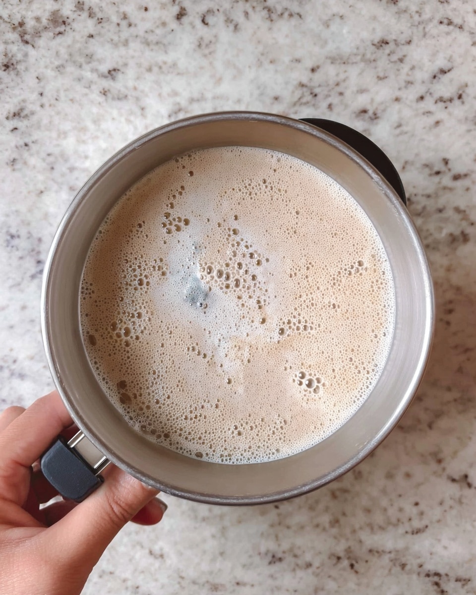 A top view of a metal mixing bowl held by a woman's hand on the bottom right. Inside the bowl is a single layer of light beige frothy liquid with many small bubbles covering the surface. The background is a white marbled textured countertop. The bowl has a black ring handle at the front edge. photo taken with an iphone --ar 4:5 --v 7