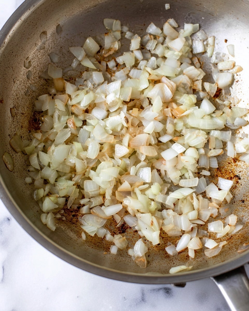 The image shows a close-up of a stainless steel pan with one layer of chopped onions that are lightly cooked and soft, with some pieces showing a pale golden brown color. The onions have a slightly translucent texture mixed with some white tones, scattered unevenly across the pan's bottom. The pan's surface has some darker browned spots, hinting at the cooking process, and the background is a white marbled texture. photo taken with an iphone --ar 4:5 --v 7