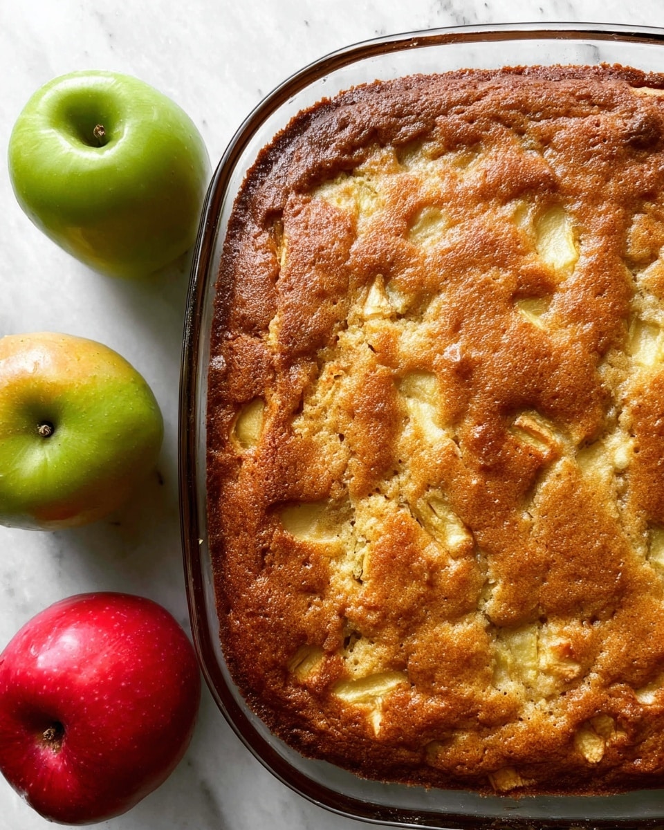 A warm apple cake in a clear glass pan with a golden brown top layer that has small chunks of apple visible inside. The cake surface is textured with slight bumps and deeper golden edges around the pan. Near the pan are two whole apples, one red and one green, placed on a white marbled background. photo taken with an iphone --ar 4:5 --v 7