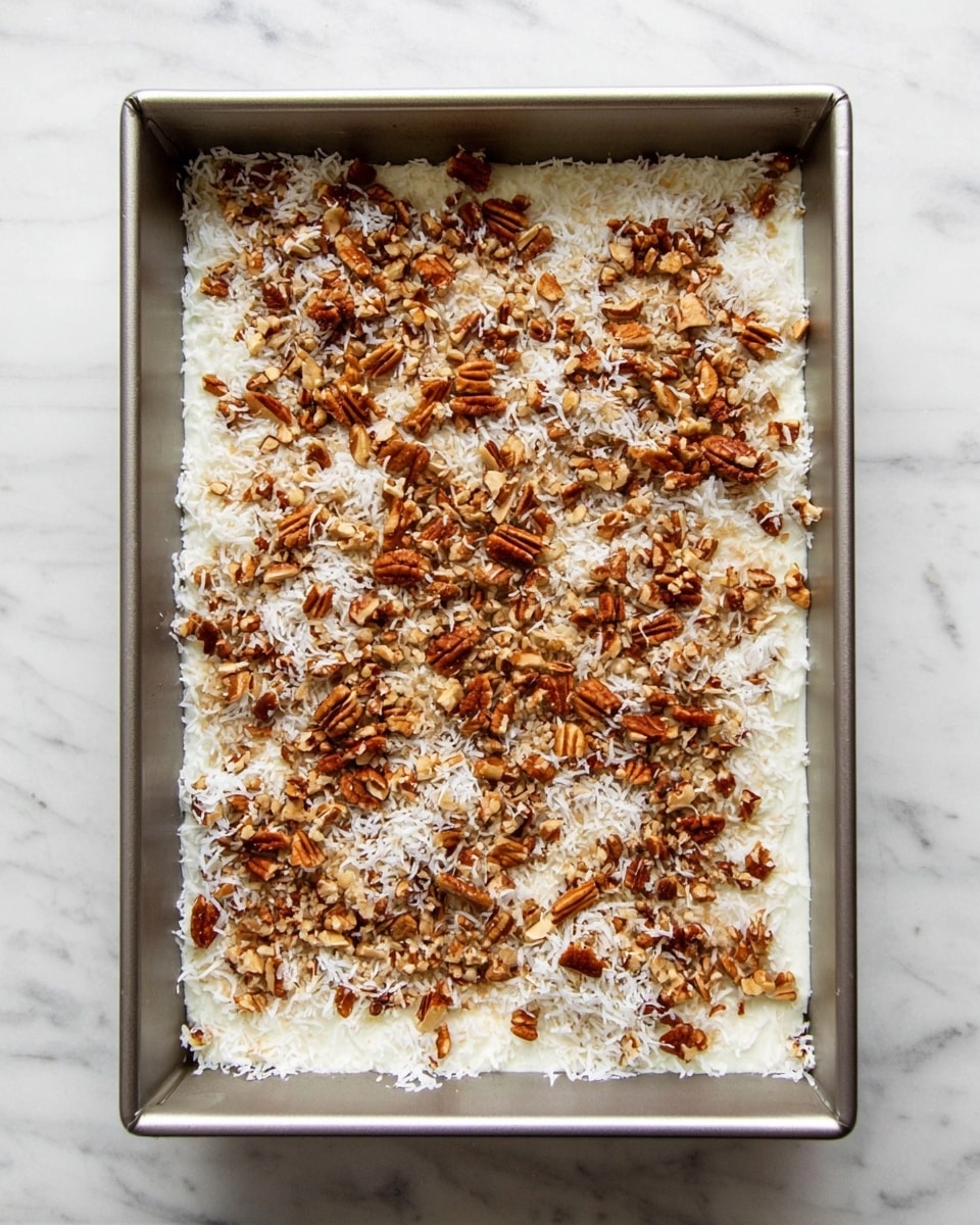 A rectangular metal baking pan holds a layered dessert with two visible layers. The bottom layer is a white creamy base, smooth and even, filling the entire pan. On top of this creamy layer is a thicker layer of shredded white coconut, spread evenly, giving a light, fibrous texture. The coconut layer is sprinkled all over with chopped pecans, varying in medium to small sizes, adding a warm brown color and a crunchy texture. The pan is placed on a white marbled surface. photo taken with an iphone --ar 4:5 --v 7