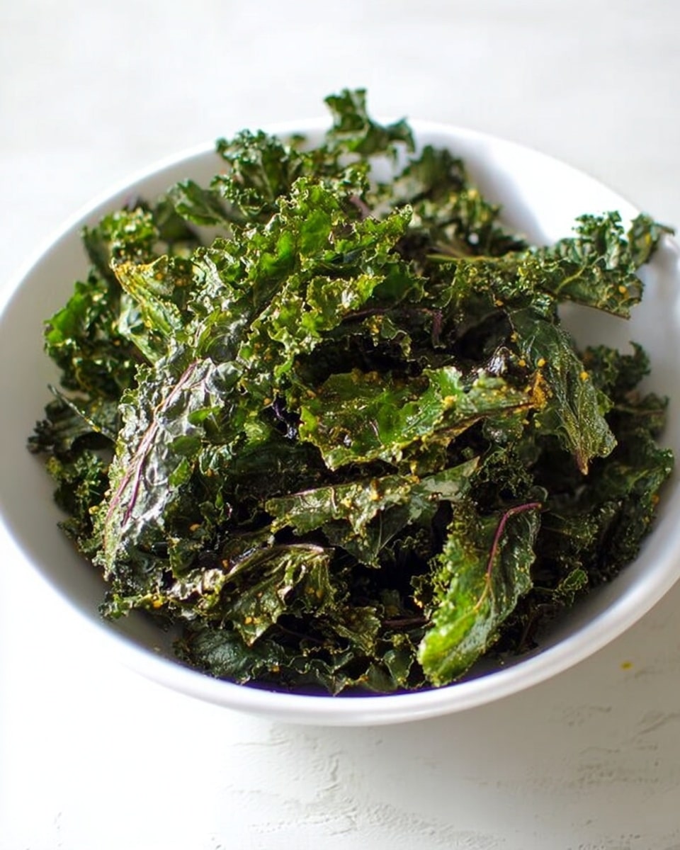 A white bowl filled with a large pile of crispy kale chips. The kale leaves are dark green with some purple stems visible, showing a rough, crunchy texture. The leaves are slightly curled and uneven with some light seasoning sprinkled on top. The bowl sits on a white marbled surface with soft natural light casting subtle shadows. Photo taken with an iphone --ar 4:5 --v 7