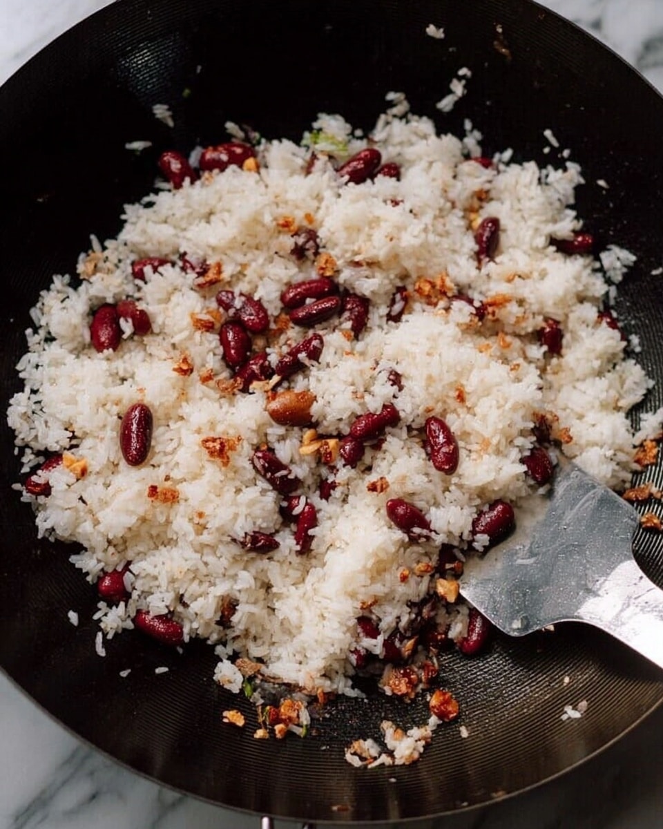 The image shows a close-up of a black wok filled with white rice mixed with small red beans and tiny brown pieces that look like nuts or seeds. The rice has a soft texture, and the beans are scattered evenly throughout. The small brown pieces add a varied texture with a crunchy look. On the right side of the wok, there is a metal spatula. The background is a white marbled surface. photo taken with an iphone --ar 4:5 --v 7