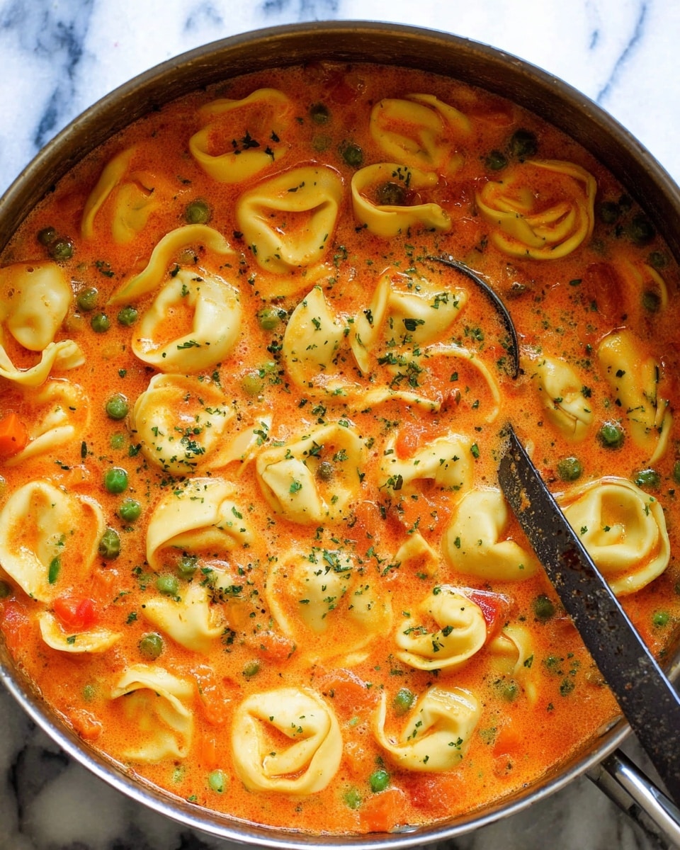 This image shows a close-up view of a large pot filled with tortellini pasta soup. The soup has a creamy tomato base with a bright orange-red color mixed with small green peas and bits of orange carrots. The tortellini noodles are pale yellow with a smooth texture and are floating on the surface in various shapes. There are tiny green herbs sprinkled over the soup, adding a touch of color contrast. A metal spoon with a dark handle is partially submerged in the soup, resting on the right side of the pot. The pot is set on a white marbled surface. photo taken with an iphone --ar 4:5 --v 7