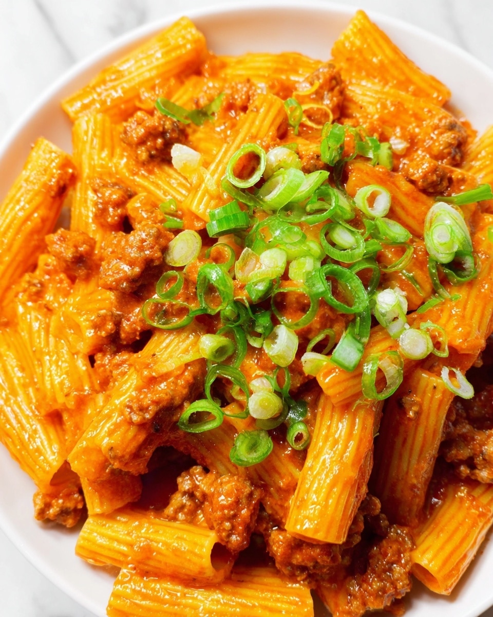 A close-up view of a white bowl filled with rigatoni pasta covered in a rich, orange-red sauce mixed with small pieces of cooked meat. On top, there is a generous layer of thinly sliced green onions, adding a bright green color and fresh texture. The rigatoni tubes have a smooth surface and are coated evenly by the sauce, sitting on a white marbled texture background. Photo taken with an iphone --ar 4:5 --v 7