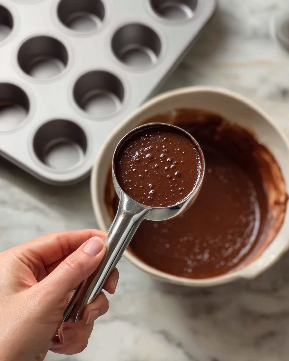 A woman's hand is holding a silver scoop filled with thick, smooth dark brown chocolate batter with small bubbles on its surface. In the background, there is a white muffin tray with empty circular cups, and a large bowl filled with more chocolate batter sits next to it. The scene is set on a white marbled surface. photo taken with an iphone --ar 4:5 --v 7