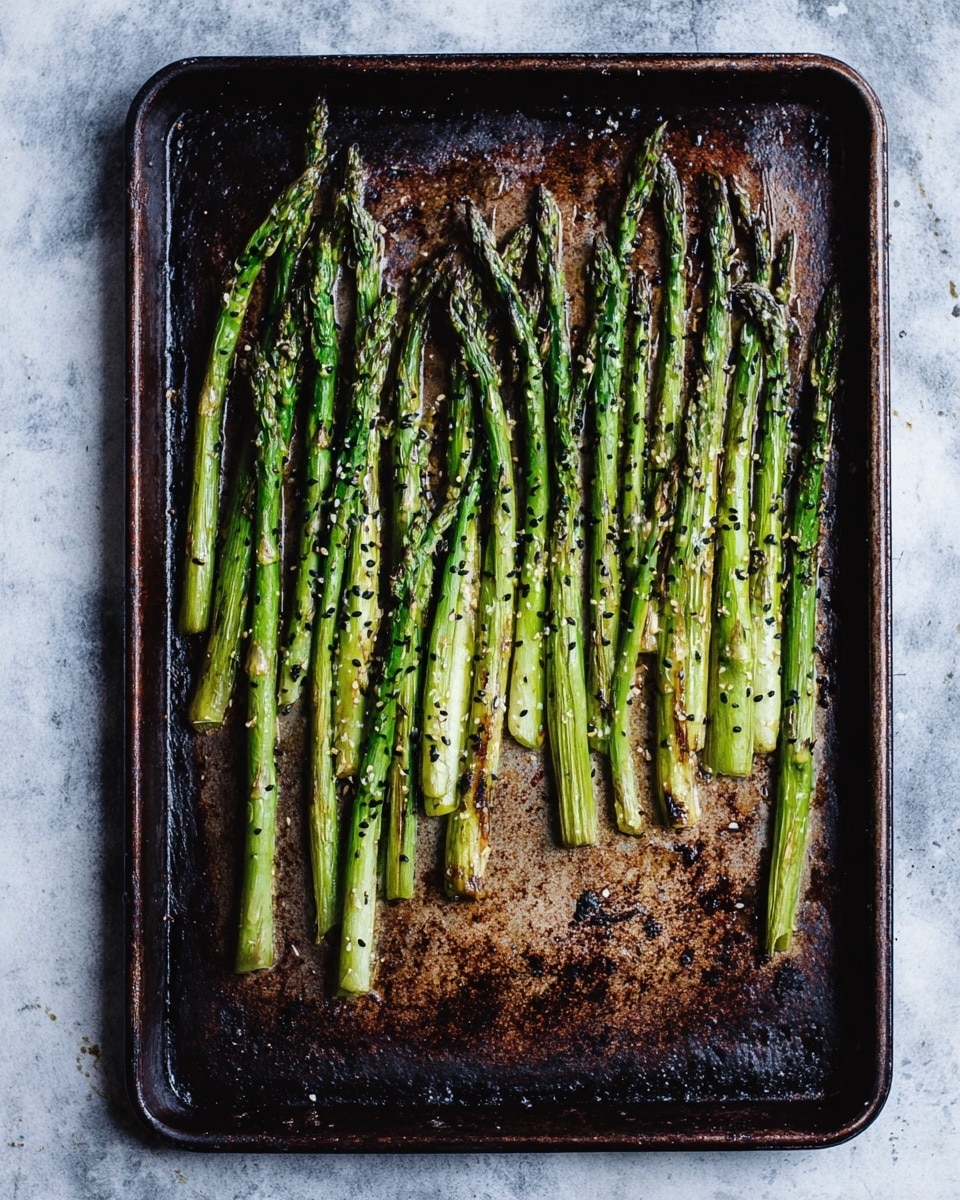 A dark, well-used metal baking tray holds a single layer of roasted asparagus spears, arranged mostly upright from left to right. The asparagus has a mix of bright to dark green shades, with some parts showing slight browning from roasting. Small black sesame seeds are sprinkled over the spears, adding little black spots on the green stalks. The textured baking tray shows charred and baked marks in rusty brown and black colors around the vegetables. The background has a white marbled texture. Photo taken with an iphone --ar 4:5 --v 7