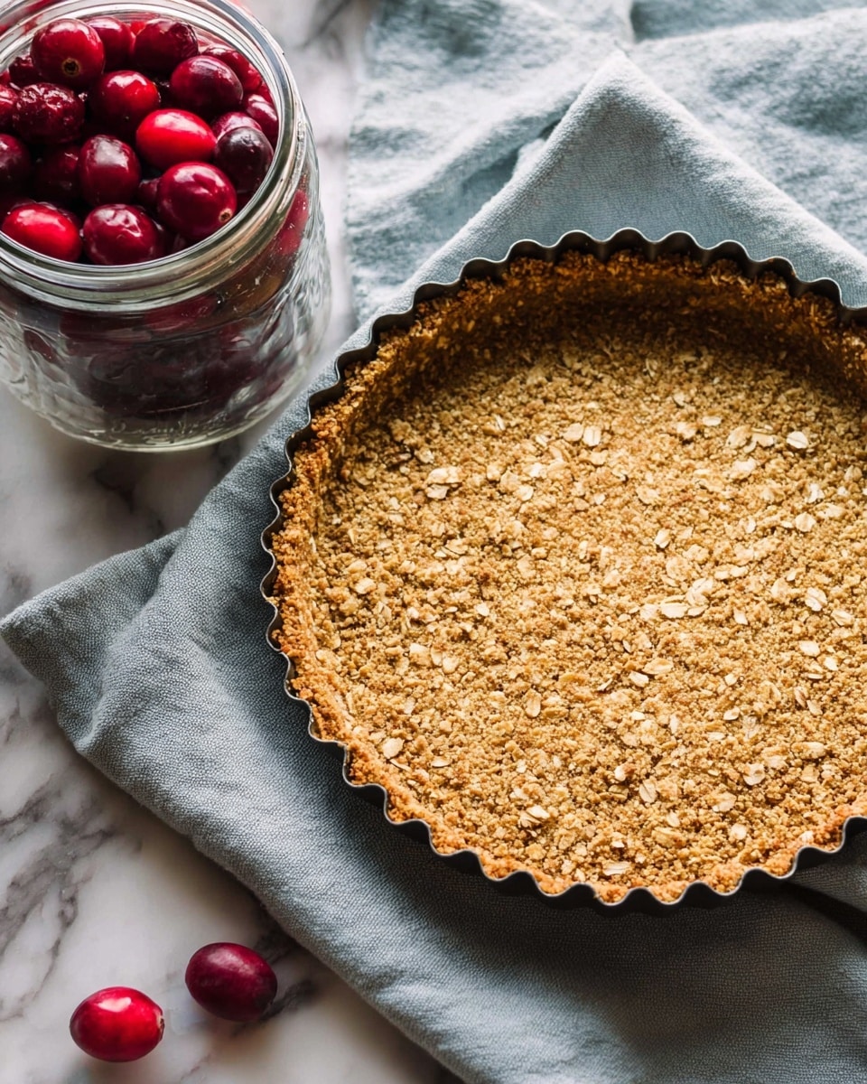 A close-up view of a round oat crust in a tart pan, showing a single textured layer of golden-brown oats pressed evenly to make the crust shape, with some areas slightly darker indicating baked spots. The crust sits on a light gray cloth with a soft, wrinkled texture, scattered with a few bright red cranberries around the pan. To the left, a clear glass jar is full of fresh, shiny red cranberries. All this rests on a white marbled surface with subtle veins and soft light. photo taken with an iphone --ar 4:5 --v 7