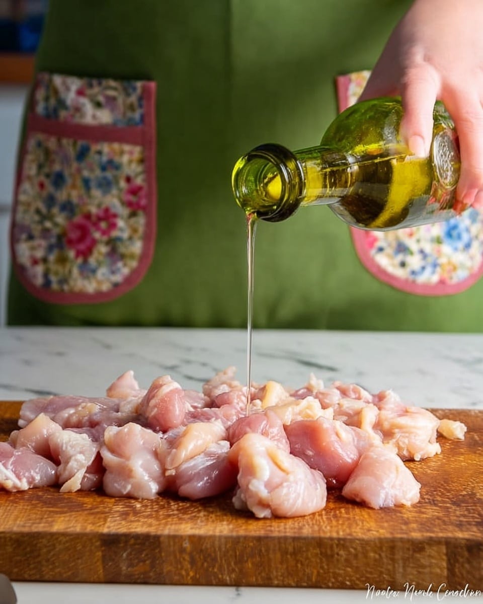 On a wooden board, there is a single layer of small raw pink chicken pieces spread out. A woman’s hand is pouring clear golden oil from a green-tinted glass bottle over the chicken. The background shows a white marbled surface below the board and part of the person wearing a green apron with two floral-patterned pockets. The lighting is soft and natural, highlighting the texture of the chicken and the oil. photo taken with an iphone --ar 4:5 --v 7