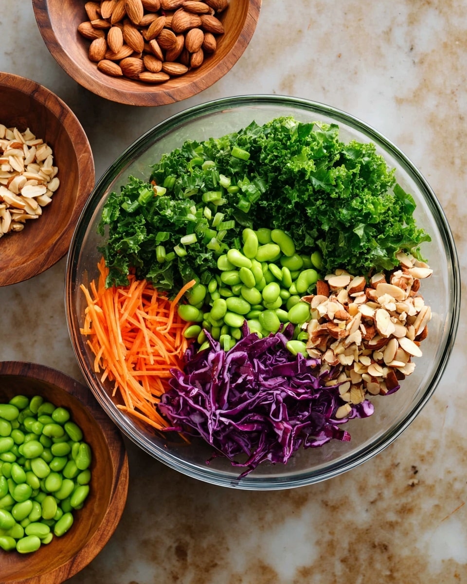A clear glass bowl holds a colorful and fresh salad with six visible layers arranged in sections: bright green chopped kale on the upper left, dark green chopped scallions below the kale, thin orange carrot sticks at the bottom left, bright green edamame beans occupying the lower right side, chopped almonds with light brown skin on the upper right and part of the middle area, and shredded purple cabbage in the middle right. Near the bowl, two wooden bowls are placed on a white marbled texture surface, one filled with chopped almonds and the other with edamame beans. Photo taken with an iphone --ar 4:5 --v 7