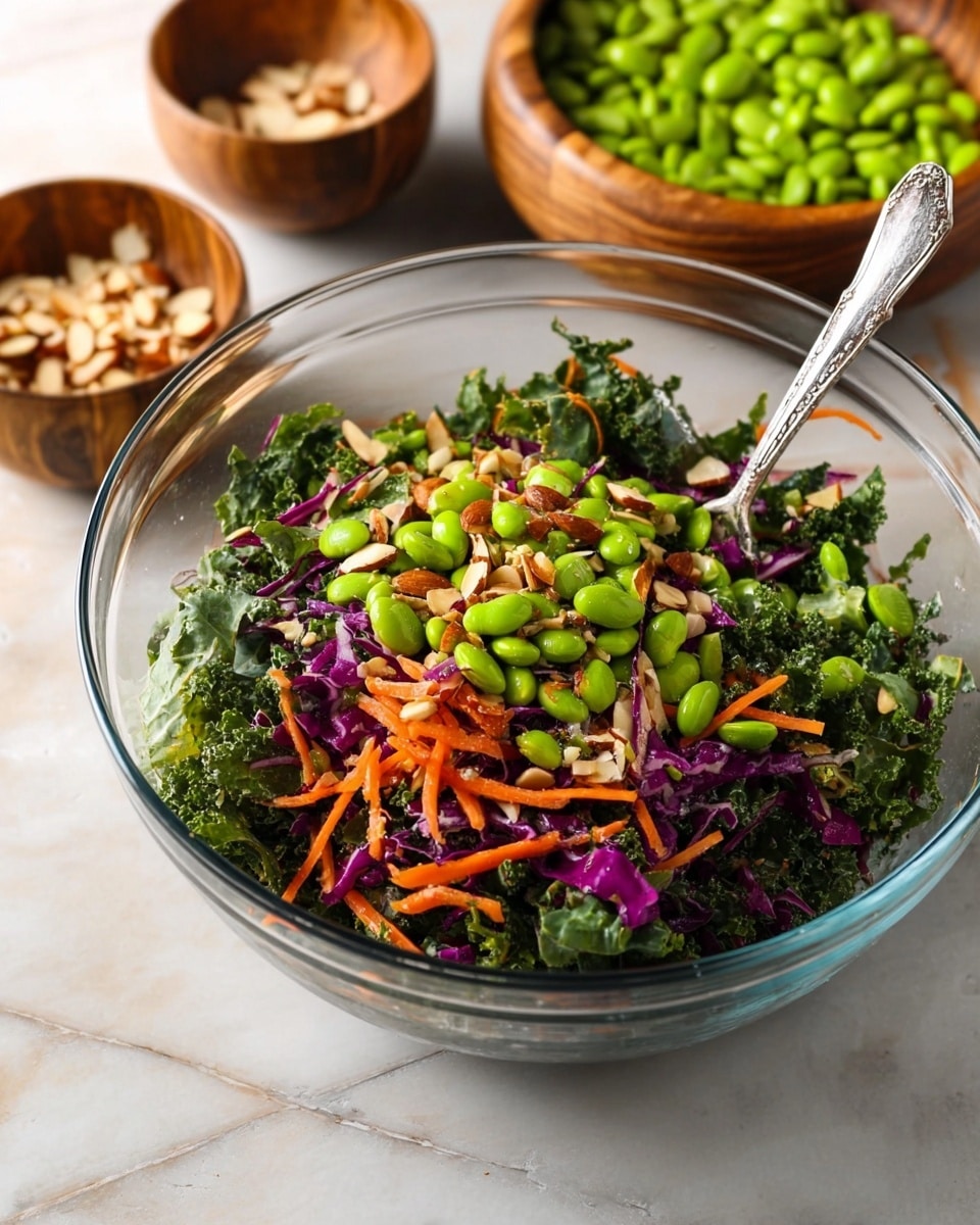 A clear glass bowl filled with a fresh salad sits on a white marbled surface. The salad has a base layer of dark green kale leaves with a rough texture. On top of the kale, there are thin, bright orange carrot strips and scattered thin strips of purple cabbage. A layer of bright green edamame beans is spread over the vegetables. Small pieces of chopped almonds are sprinkled throughout the salad, adding a light brown color and crunchy texture. A detailed silver spoon rests inside the bowl, partly submerged in the salad. Two small wooden bowls are in the background, one with more green edamame beans and the other with chopped almonds. Photo taken with an iphone --ar 4:5 --v 7