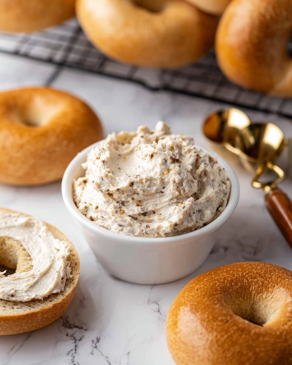 The image shows a close-up of a toasted bagel sandwich stacked on another whole toasted bagel. The top bagel has a golden-brown, slightly textured surface, and inside is a creamy, white spread with a thick, slightly chunky texture. Behind this stack, there are more toasted bagels inside a black wire basket. The foreground also has a white bowl filled with the same creamy spread, and a blue and white checkered cloth is partially visible on the left side. All items rest on a white marbled surface. Photo taken with an iphone --ar 4:5 --v 7