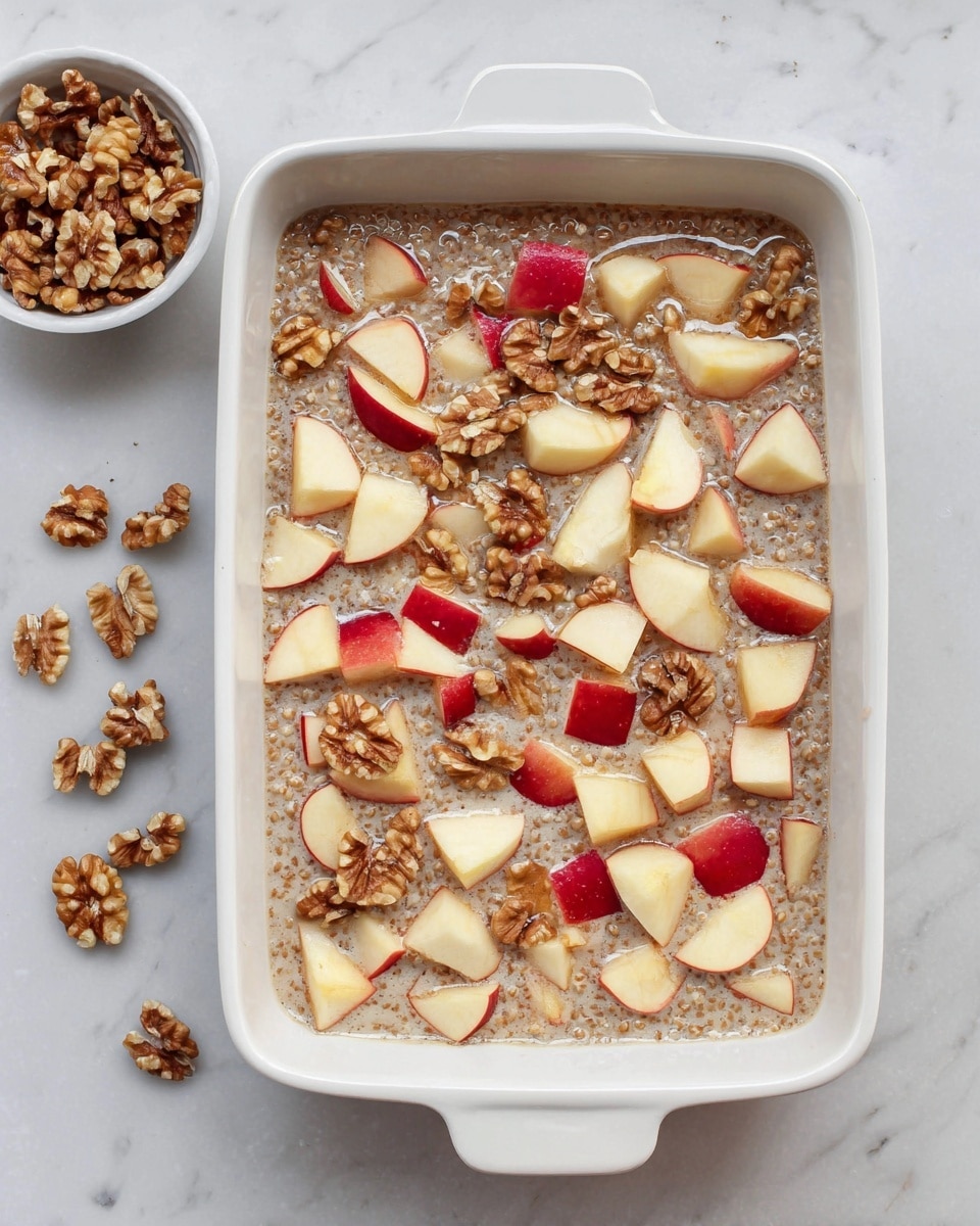 A white rectangular baking dish filled with a mixture that has small round white grains suspended in a light brown creamy liquid, topped with many small cubes of red-skinned apples scattered evenly, and several whole walnut pieces spread on top. To the top left side next to the dish, there is a small white bowl filled with more walnut pieces. The whole setup rests on a white marbled surface photo taken with an iphone --ar 4:5 --v 7